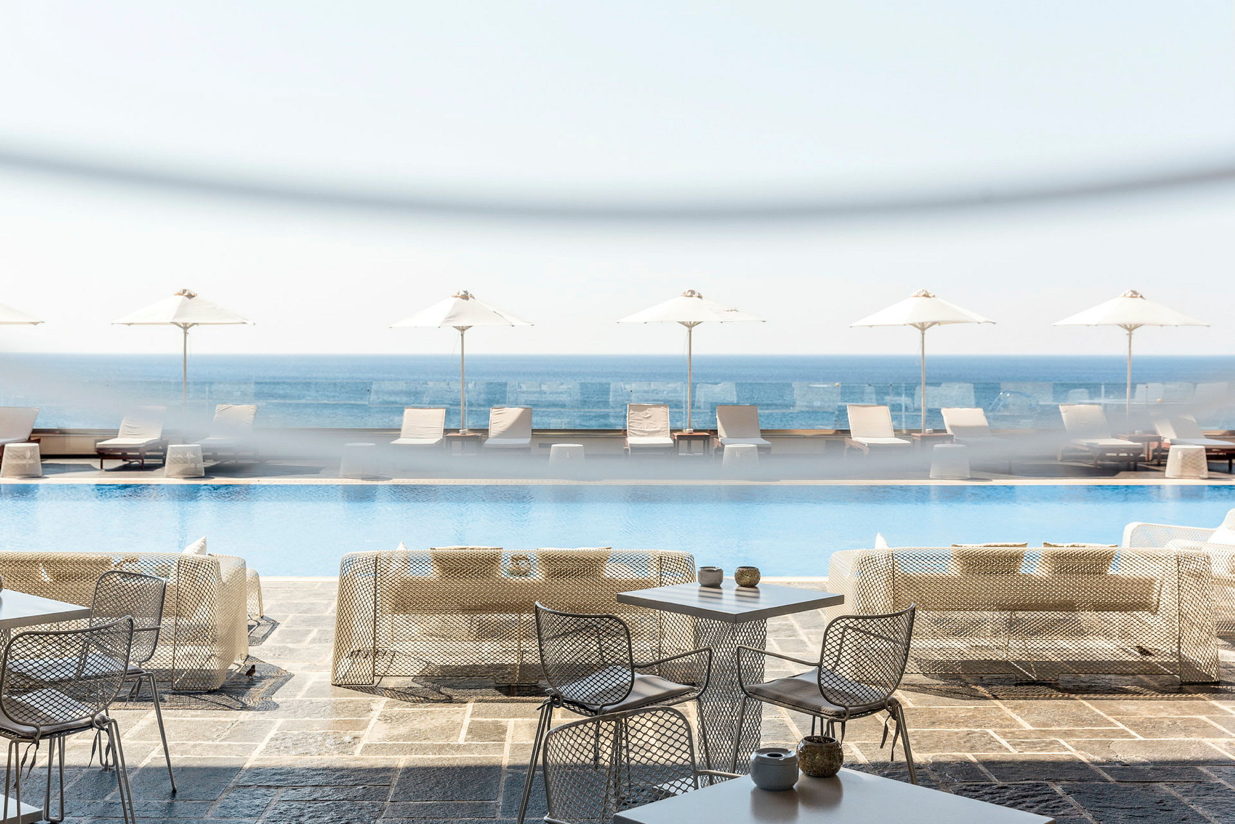 Udendørs borde og stole foran en pool, solsenge og parasoller med havet i baggrunden. (AI-genereret tekst)