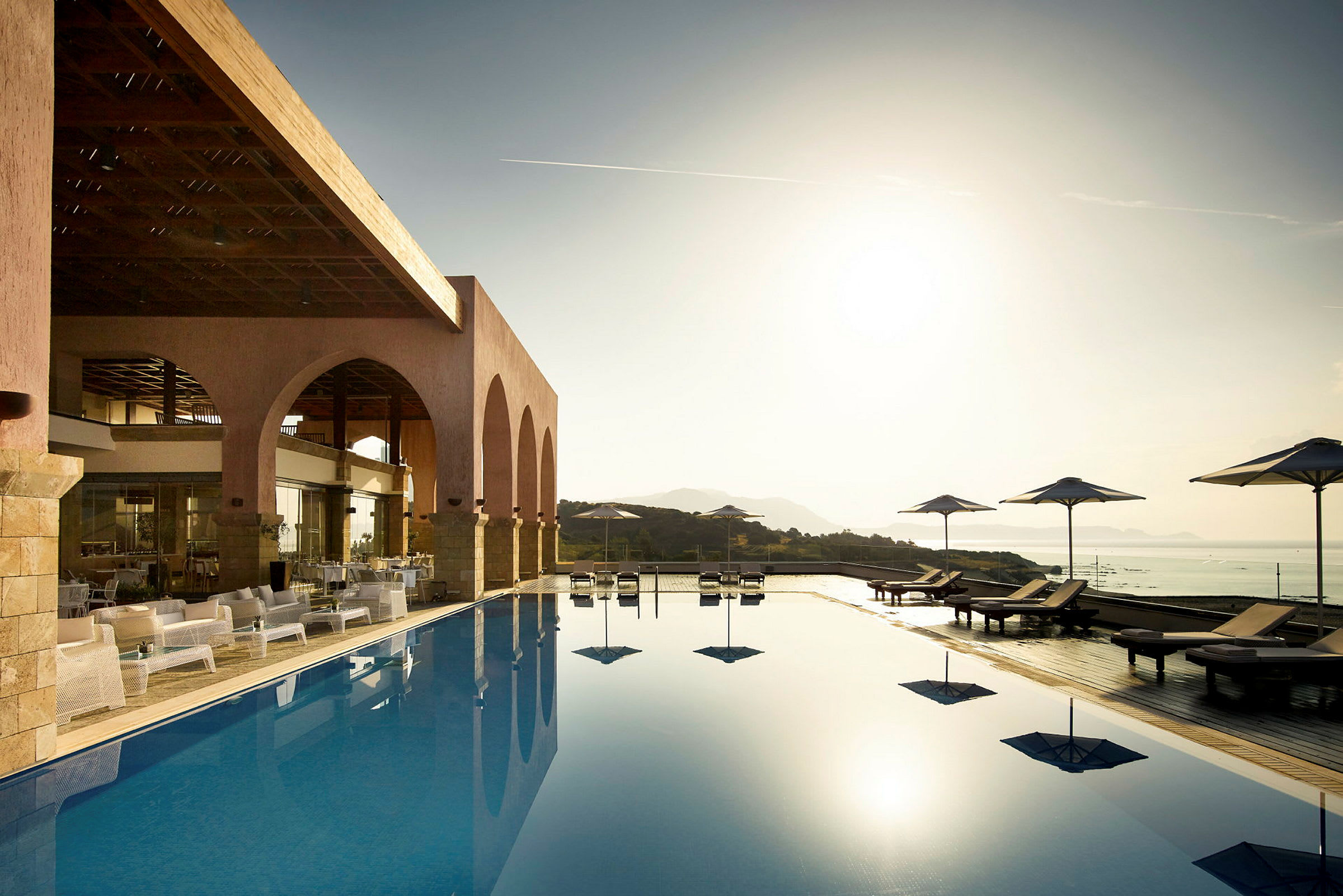 En pool med liggestole og parasoller nær en terrasse med buede åbninger. Solen går ned over landskabet. (AI-genereret tekst)