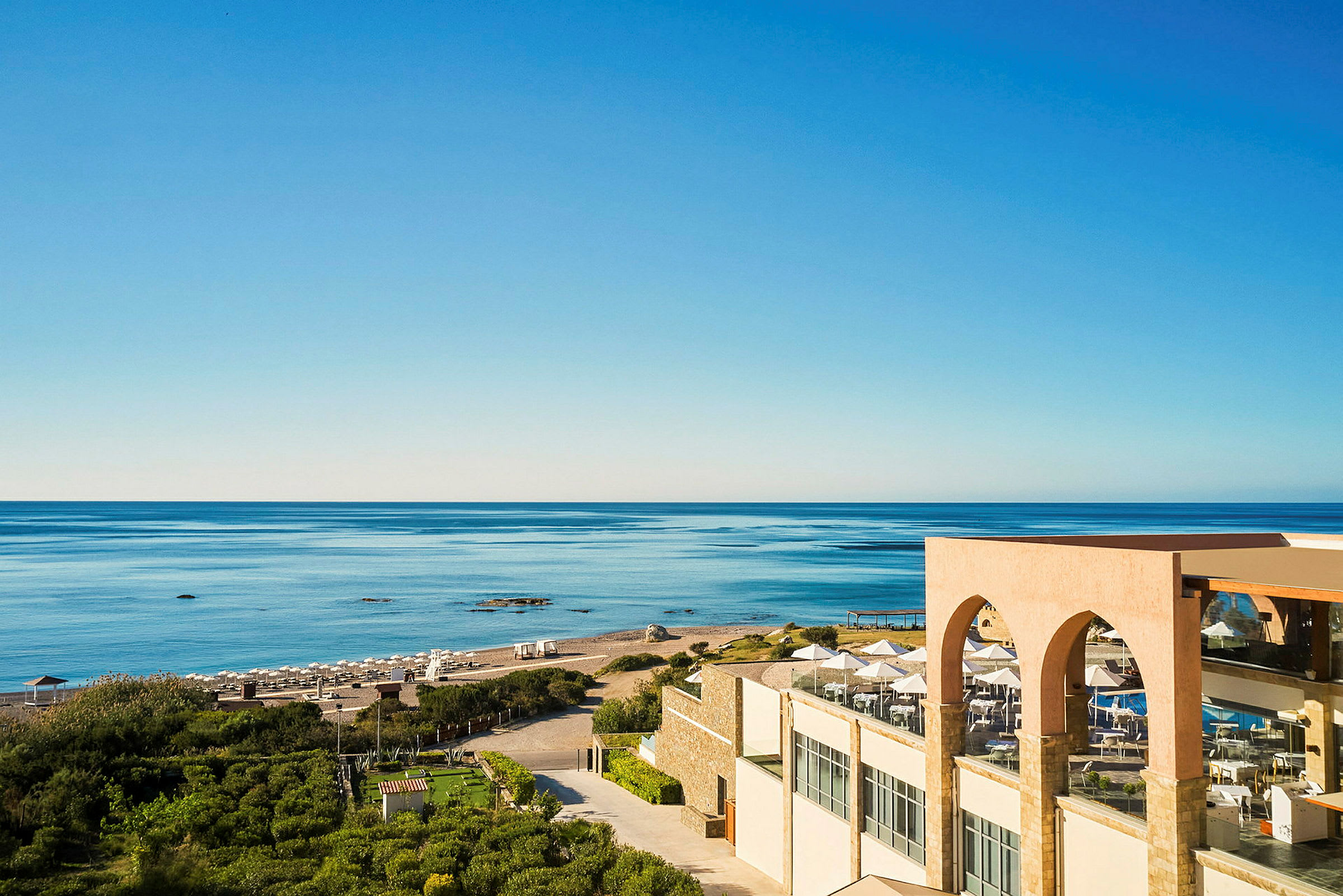Udsigt over en sandstrand, havet og et hotel med terrasse i forgrunden. (AI-genereret tekst)