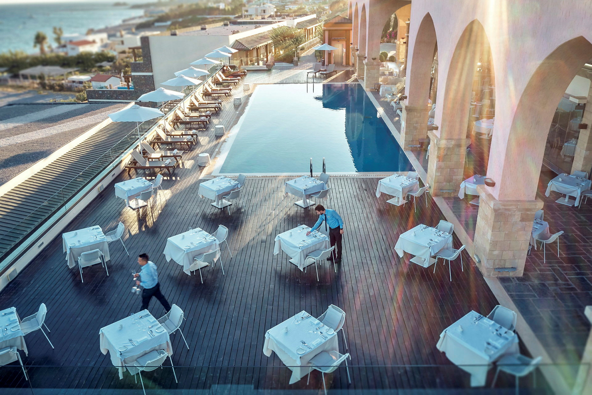 Udendørsrestaurant ved en pool med opstillede borde og stole, tjener forbereder borddækning. (AI-genereret tekst)