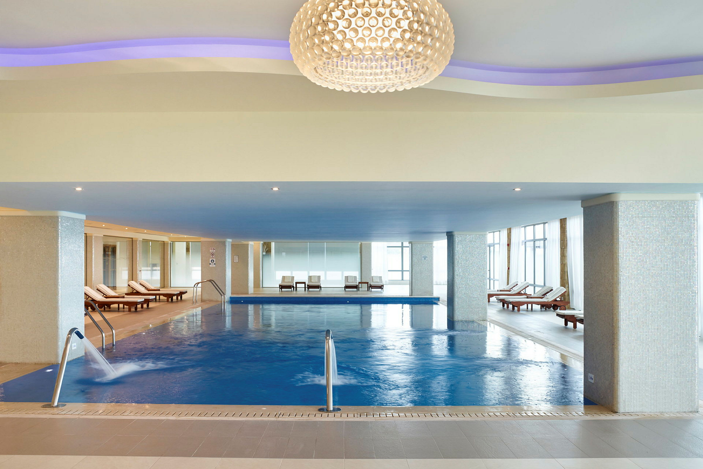 Indendørs swimmingpool med liggestole og dekorativ belysning i loftet. (AI-genereret tekst)