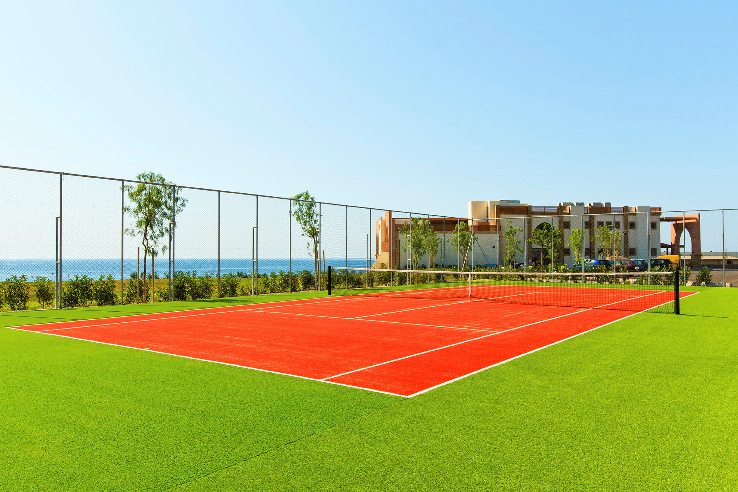 En tennisbane med rødt belægning omgivet af grønt græs, med havet og en bygning i baggrunden. (AI-genereret tekst)