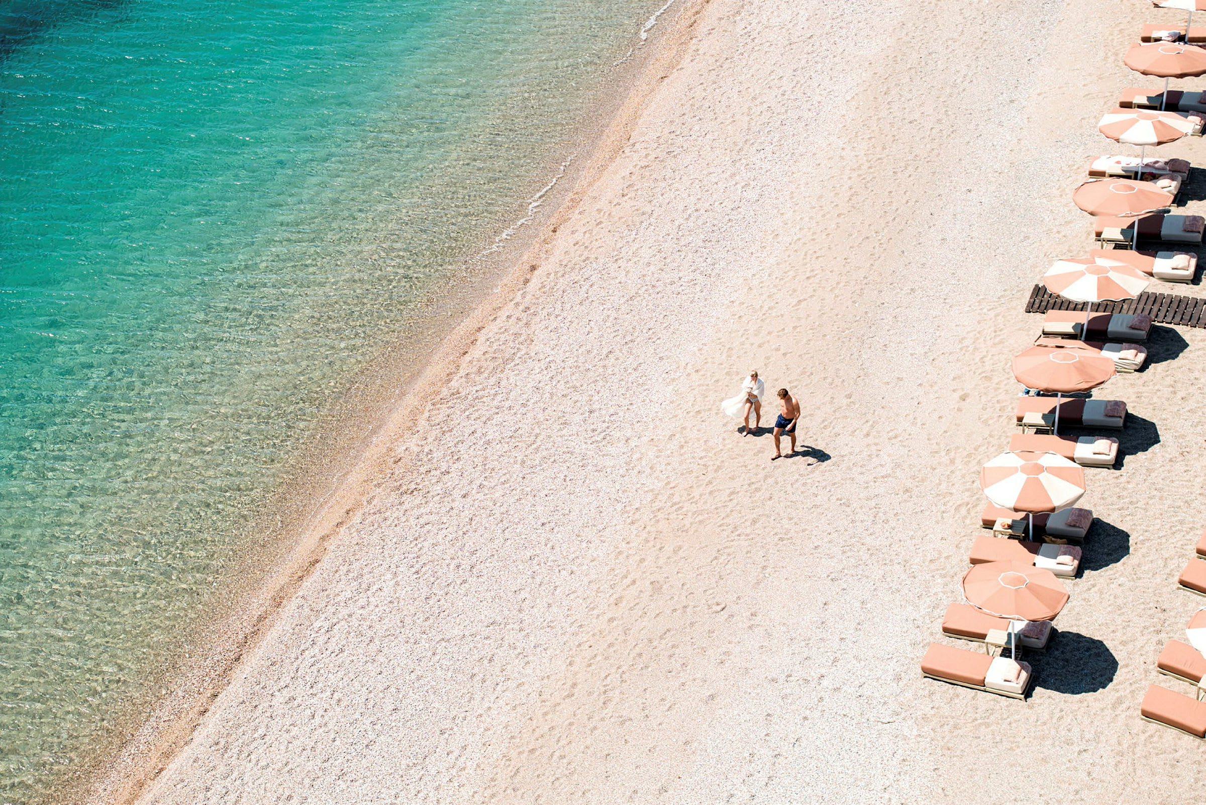 En strand med klart vand og parasoller langs den ene side. To personer går på sanden. (AI-genereret tekst)