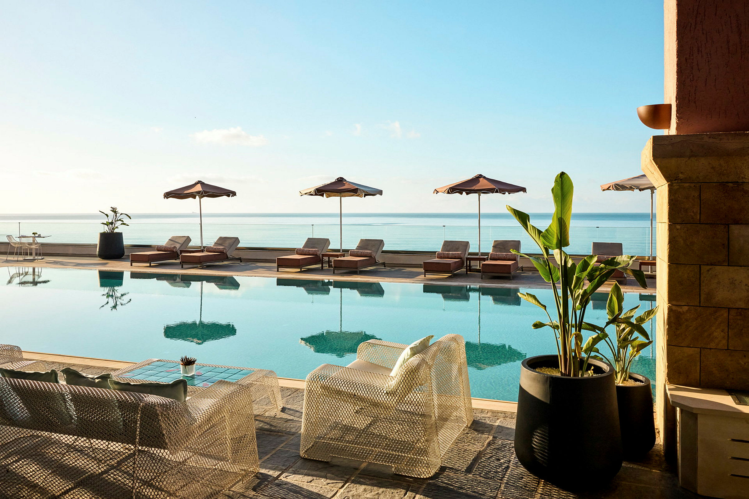En swimmingpool med liggestole og parasoller ved siden og udsigt over havet. Foran poolen står stole og borde samt store krukkeplanter. (AI-genereret tekst)