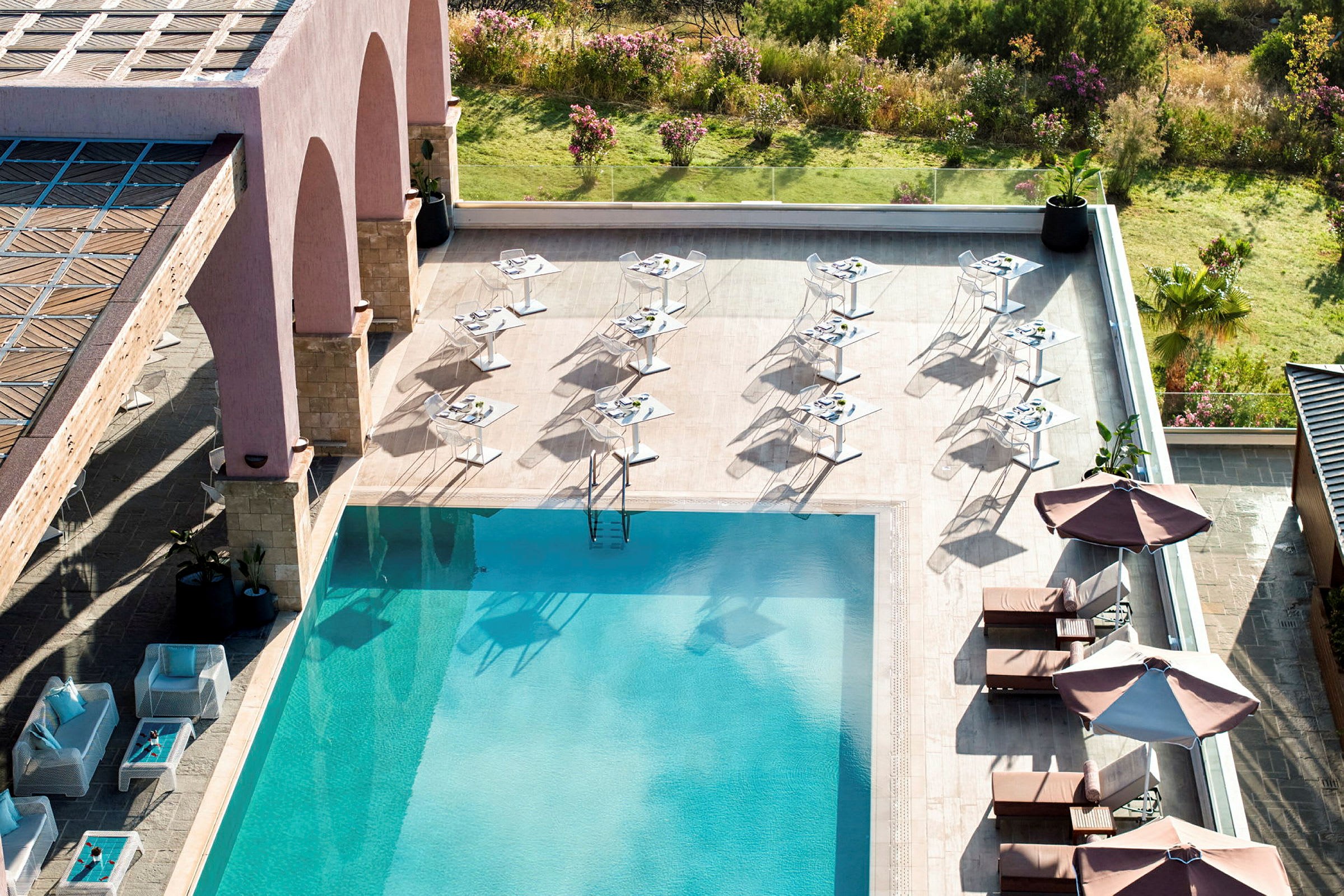 En udendørs pool med liggestole og parasoller ved siden, omgivet af borde og stole på en terrasse. (AI-genereret tekst)