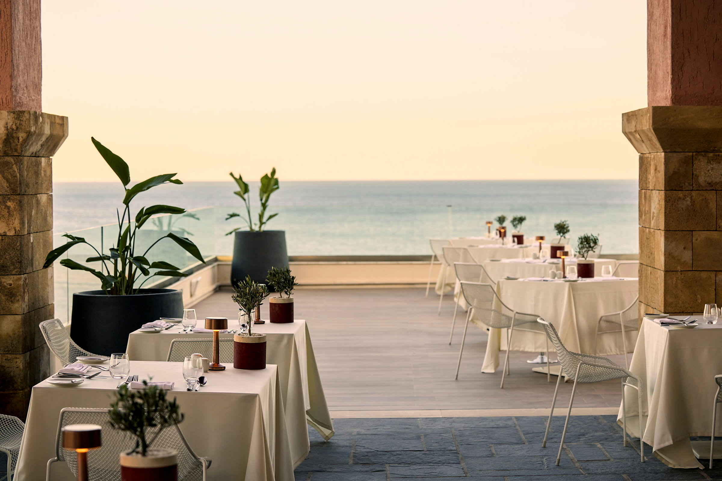Udendørs restaurant med dækkede borde, stole og dekorative planter tæt på havet. (AI-genereret tekst)
