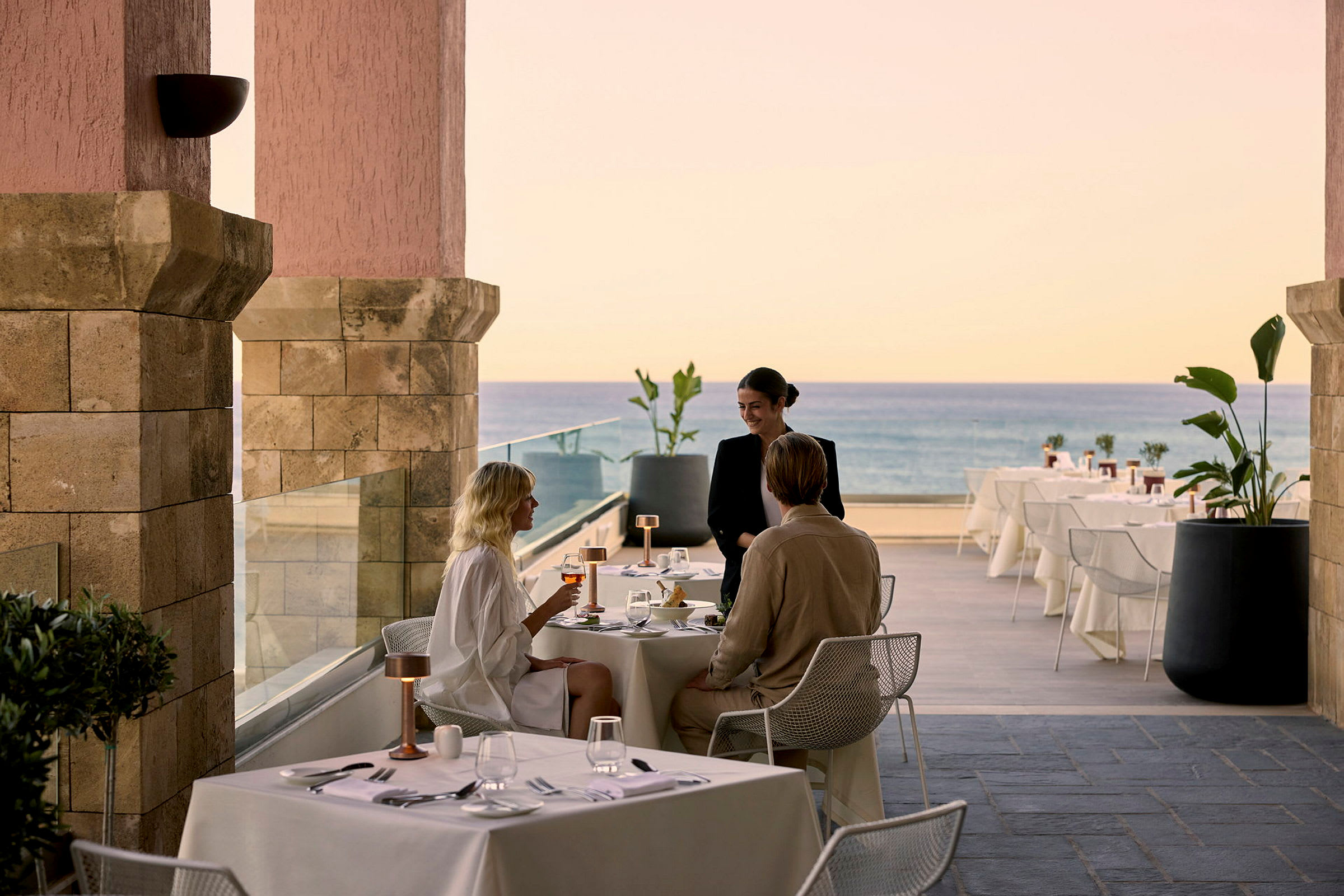 Personer, der sidder ved et dækket restaurantbord med udsigt over havet. (AI-genereret tekst)