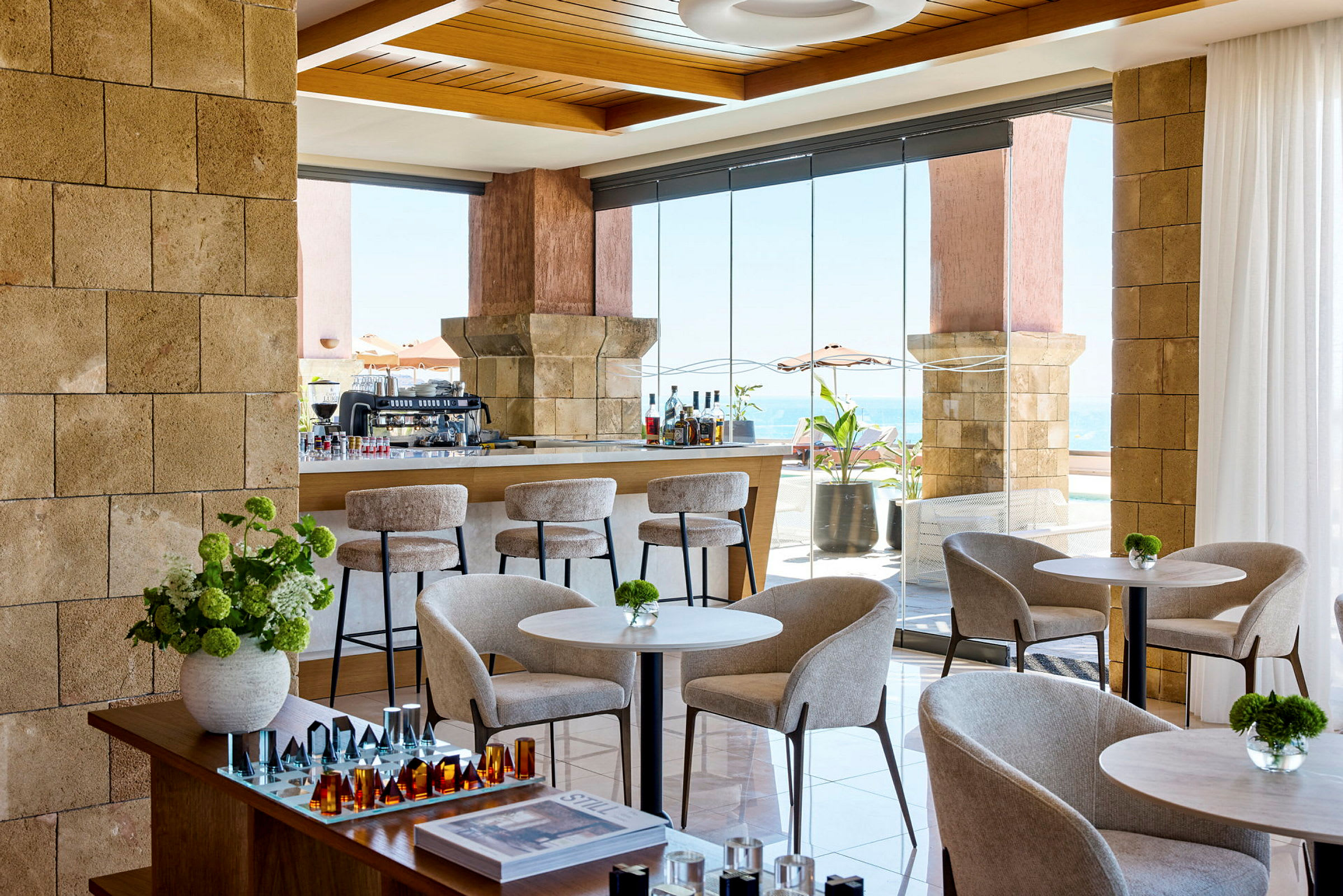 Billede af en moderne og lys lounge med siddepladser, borde og dekorative elementer. Vinduer viser udsigt mod havet. (AI-genereret tekst)
