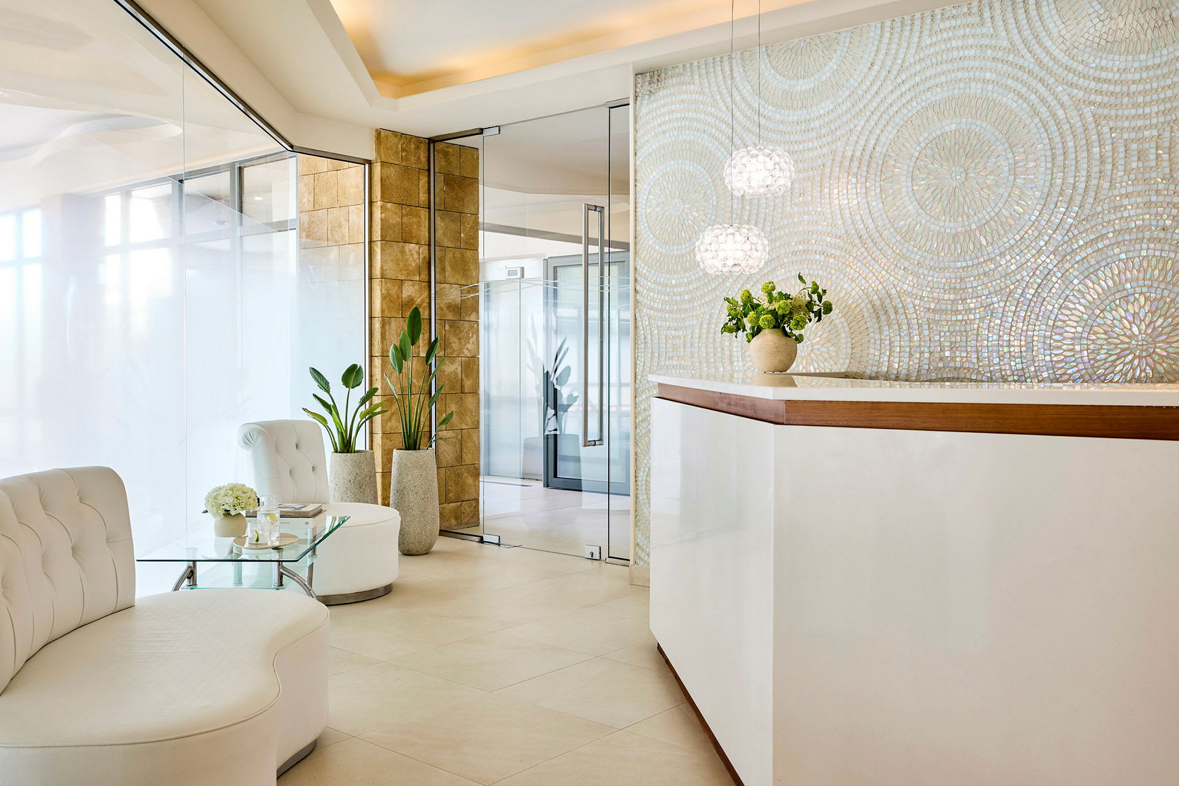 Et moderne receptionsområde med hvide møbler, glasbord og grønne planter. En stilfuld væg med iriserende mosaik findes bag skranken. (AI-genereret tekst)