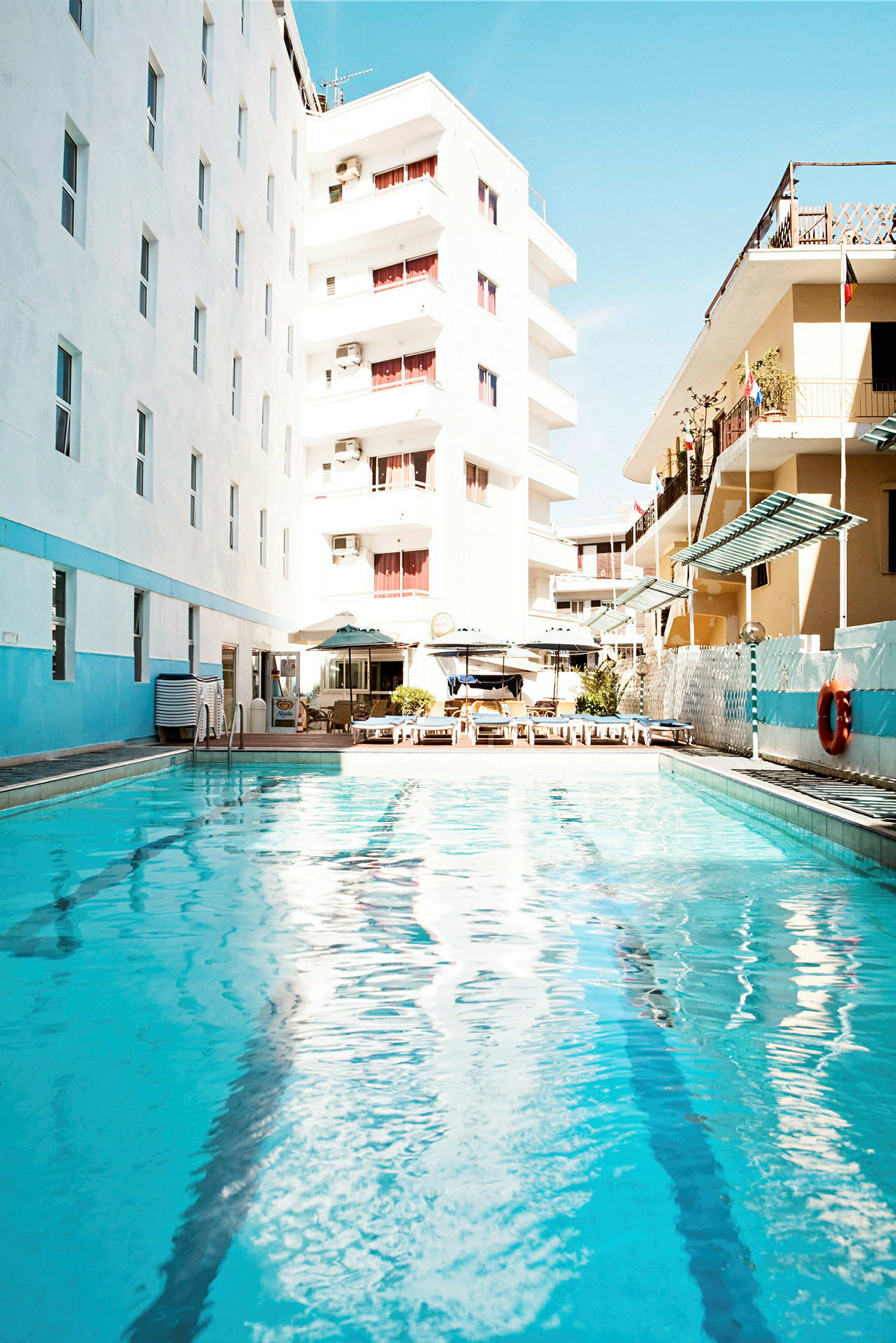 En udendørs pool omgivet af hotelbygninger og liggestole. (AI-genereret tekst)