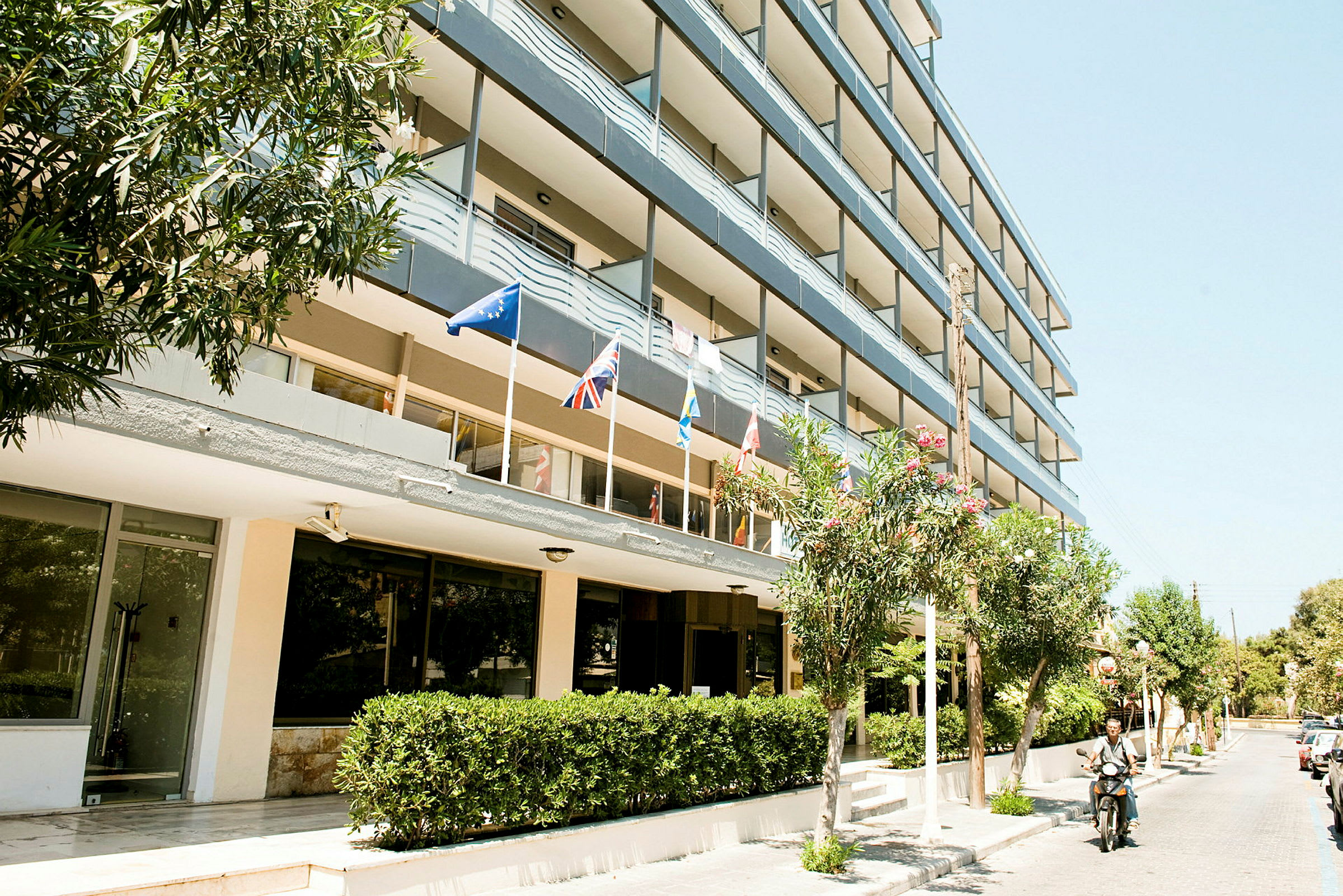 Billede af et fleretagers hotel med flag ved indgangen. (AI-genereret tekst)