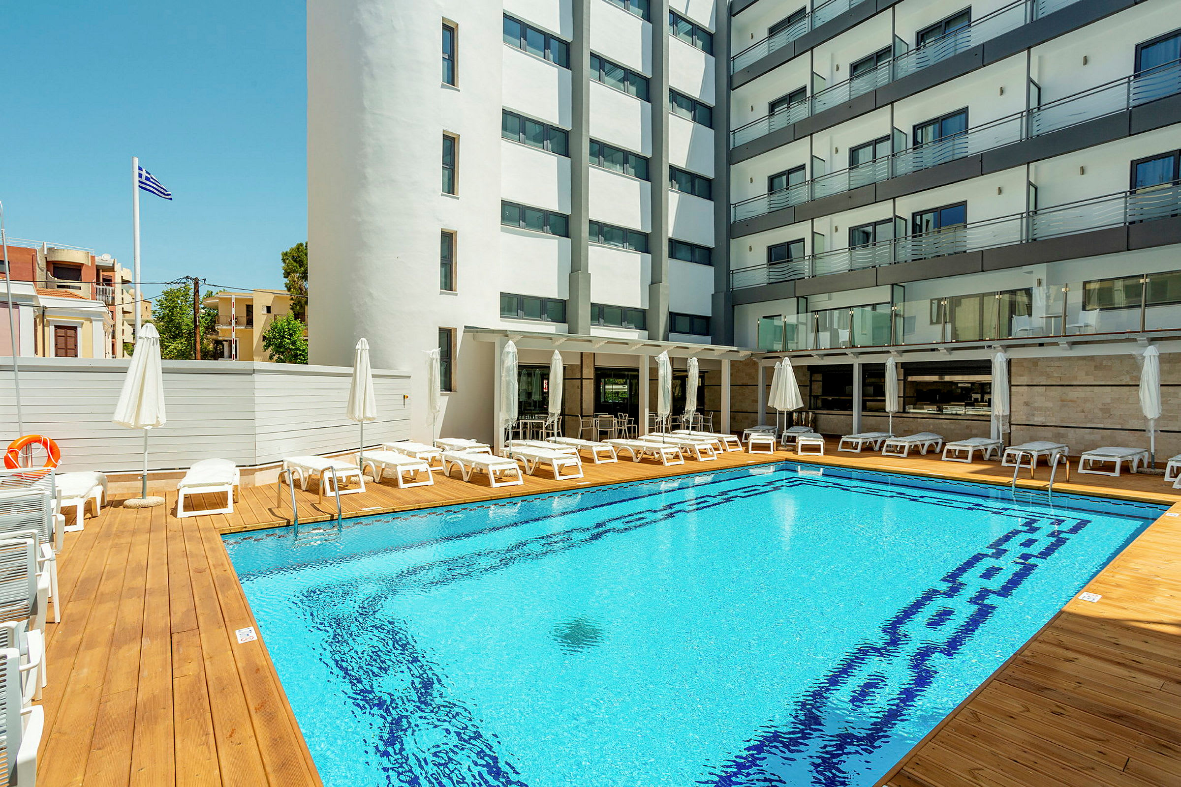 Hotelpool med liggestole og parasoller, omgivet af bygninger. (AI-genereret tekst)