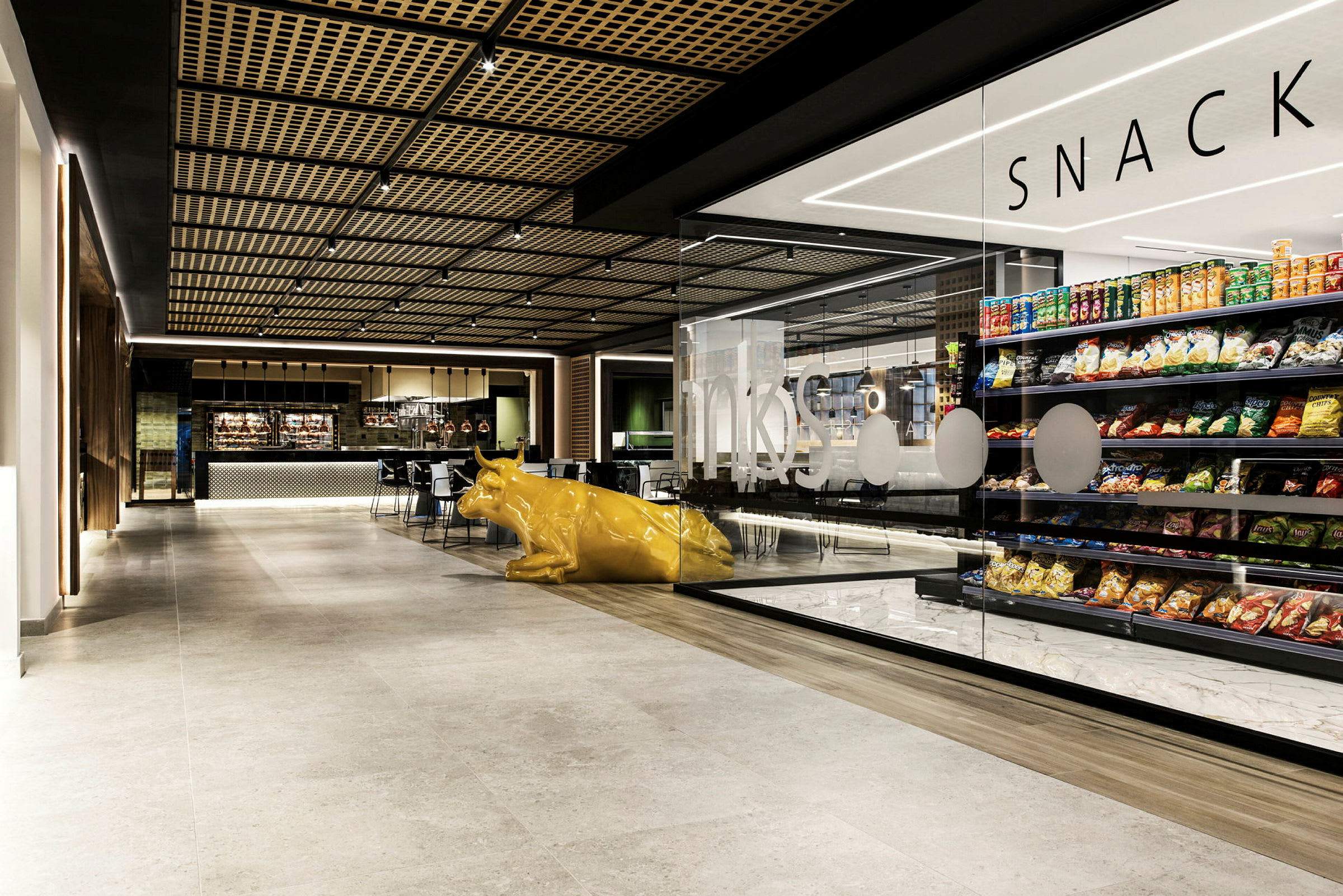 Et indendørsbillede af en moderne café med butiksreoler fyldt med snacks og en gul kofigur i forgrunden. (AI-genereret tekst)