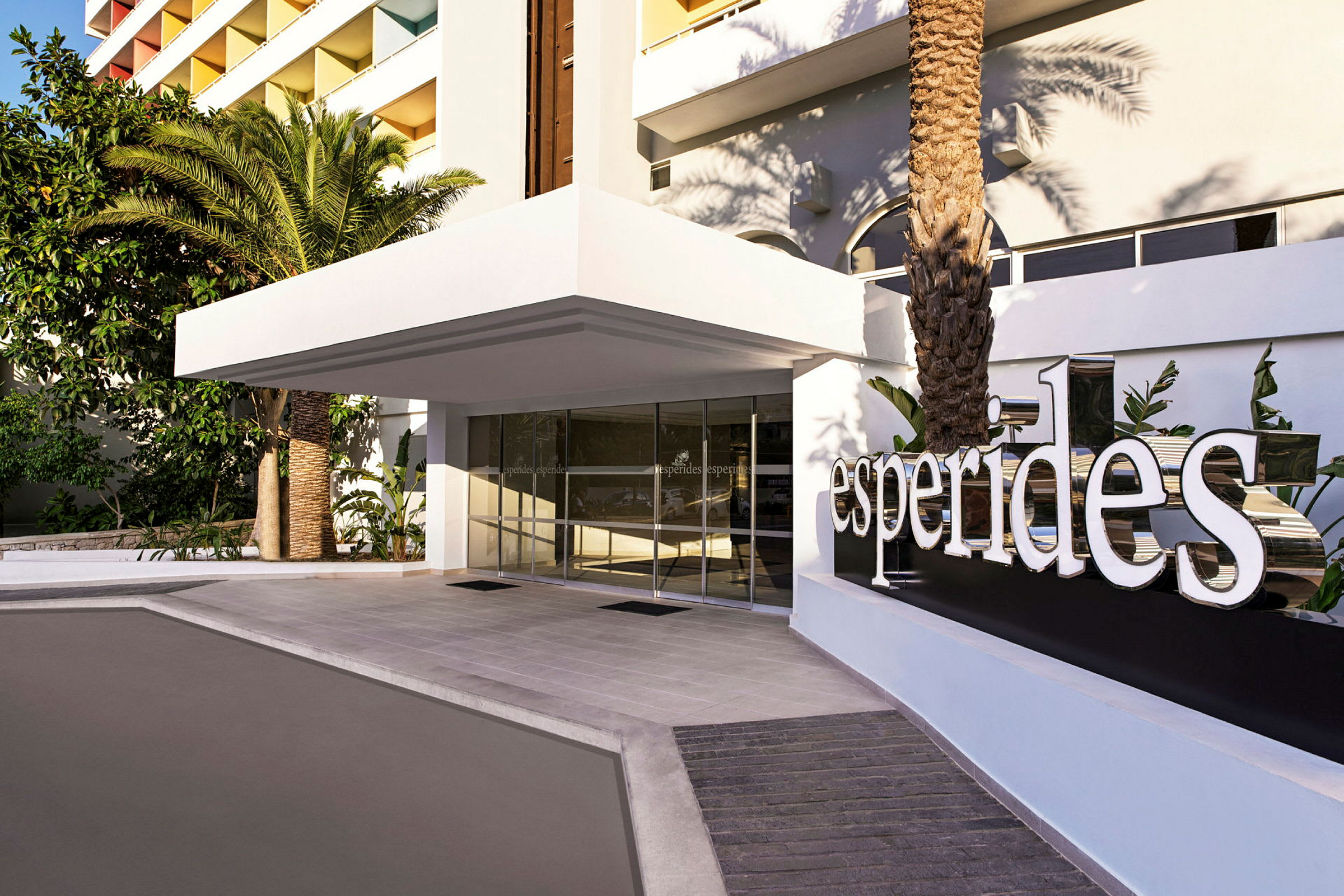 Hotelindgang med hvidt skilt, der viser teksten "Esperides" og omkringliggende grøntområde. (AI-genereret tekst)
