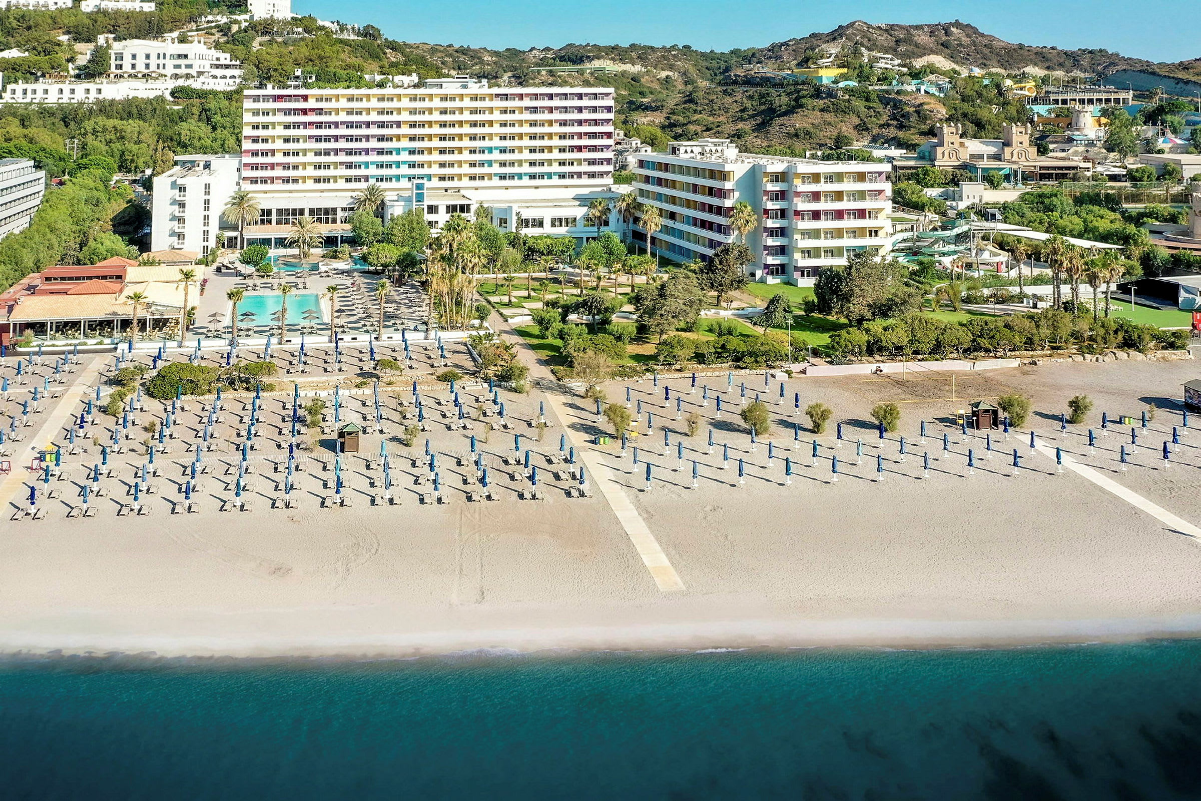 Hotelkompleks ved stranden med liggestole og parasoller og havet i forgrunden. (AI-genereret tekst)
