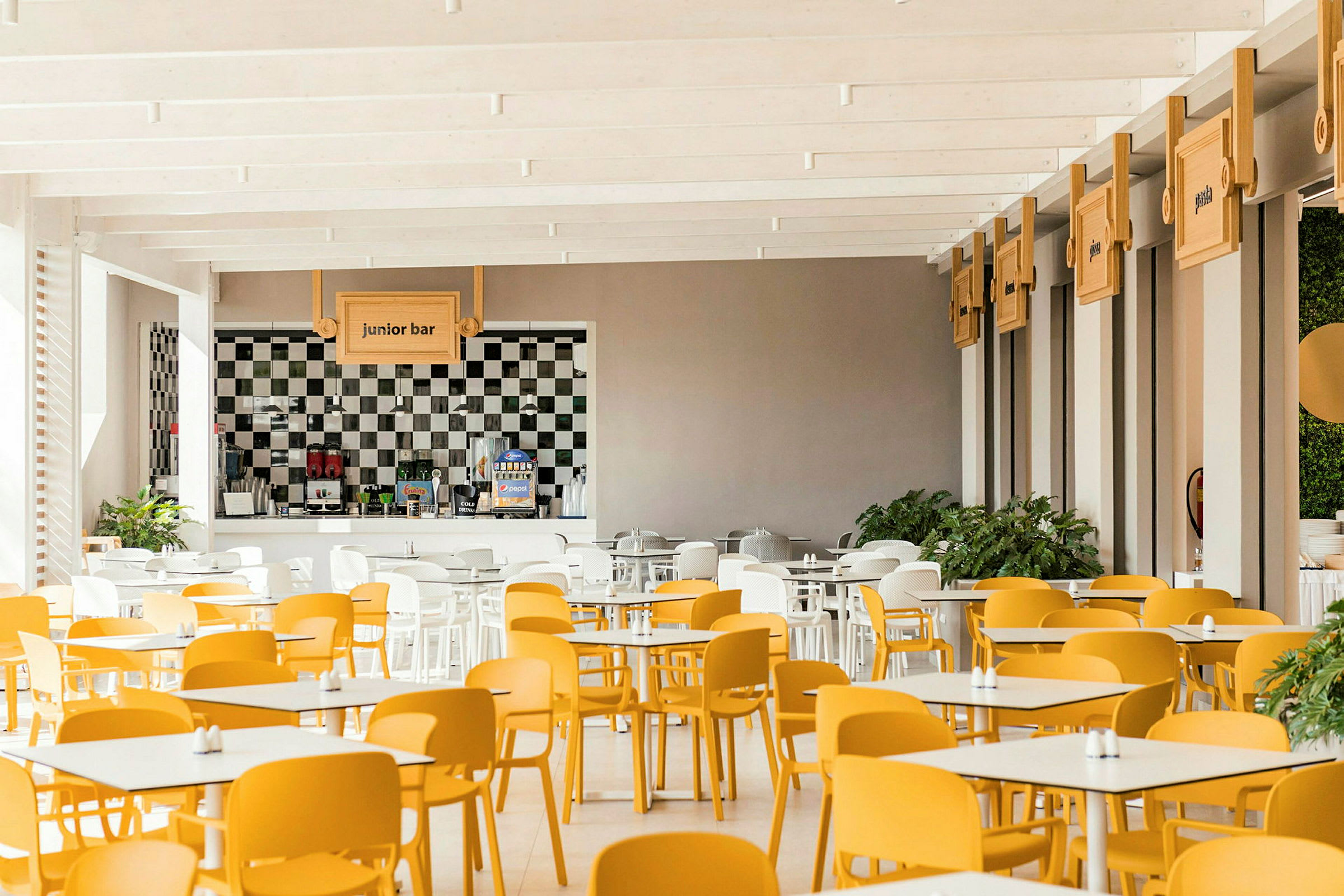 En lys spisesal med gule og hvide stole samt bordopstilling. En bardisk med skiltet "junior bar" ses i baggrunden. (AI-genereret tekst)