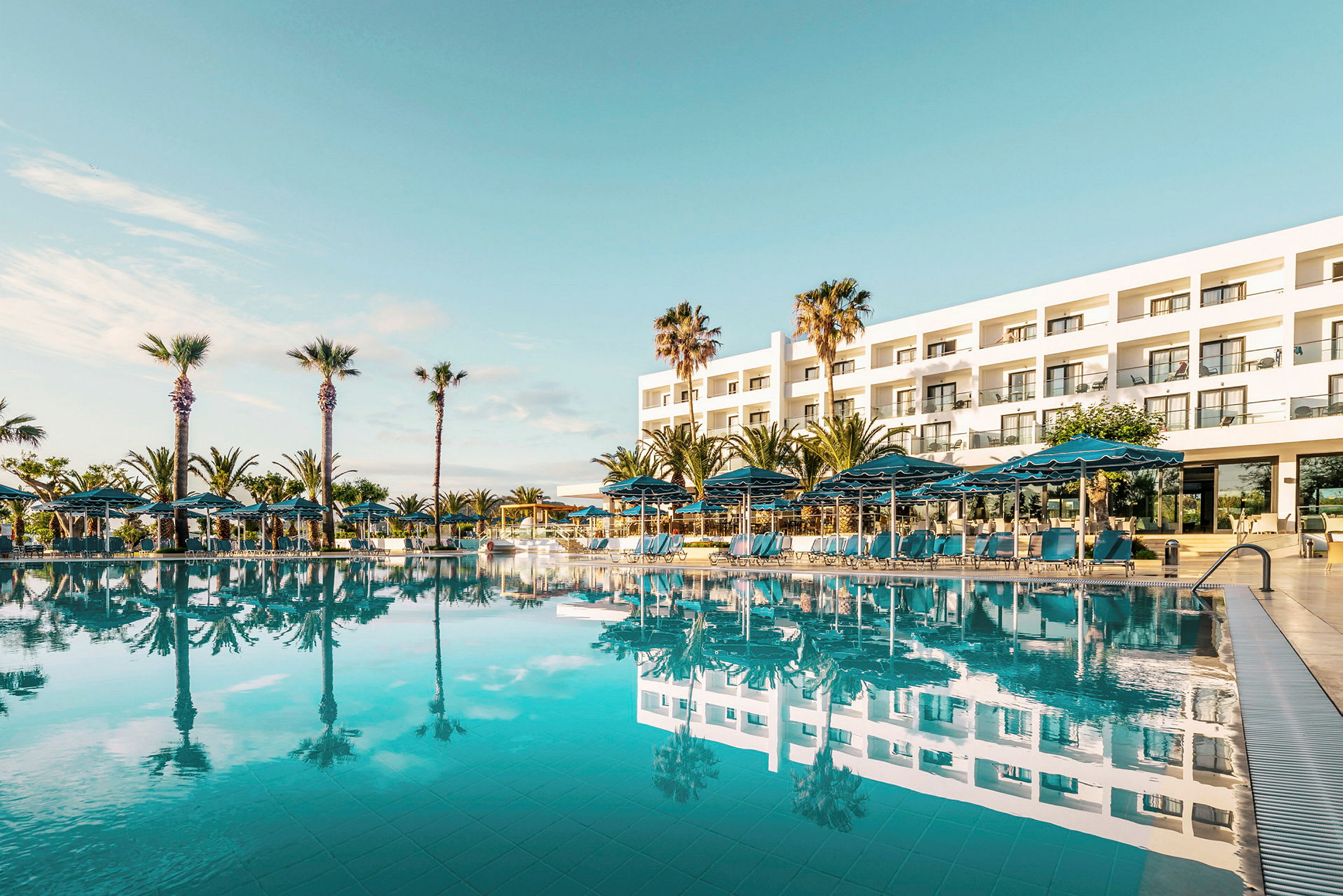 En pool med liggestole og parasoller foran et moderne hotel med palmer. (AI-genereret tekst)