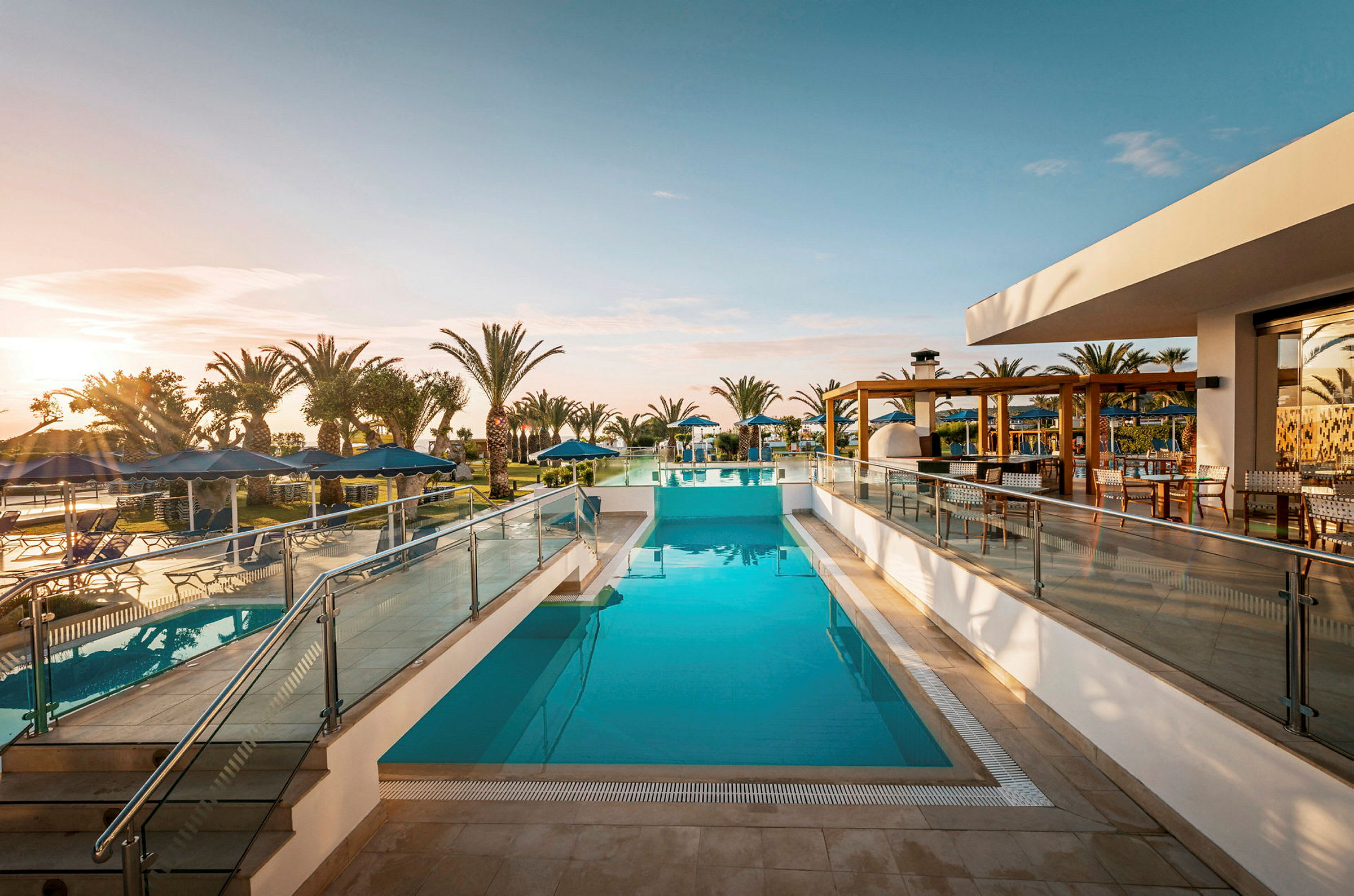 En swimmingpool ved et hotel, omgivet af palmer og solsenge under en klar himmel. (AI-genereret tekst)