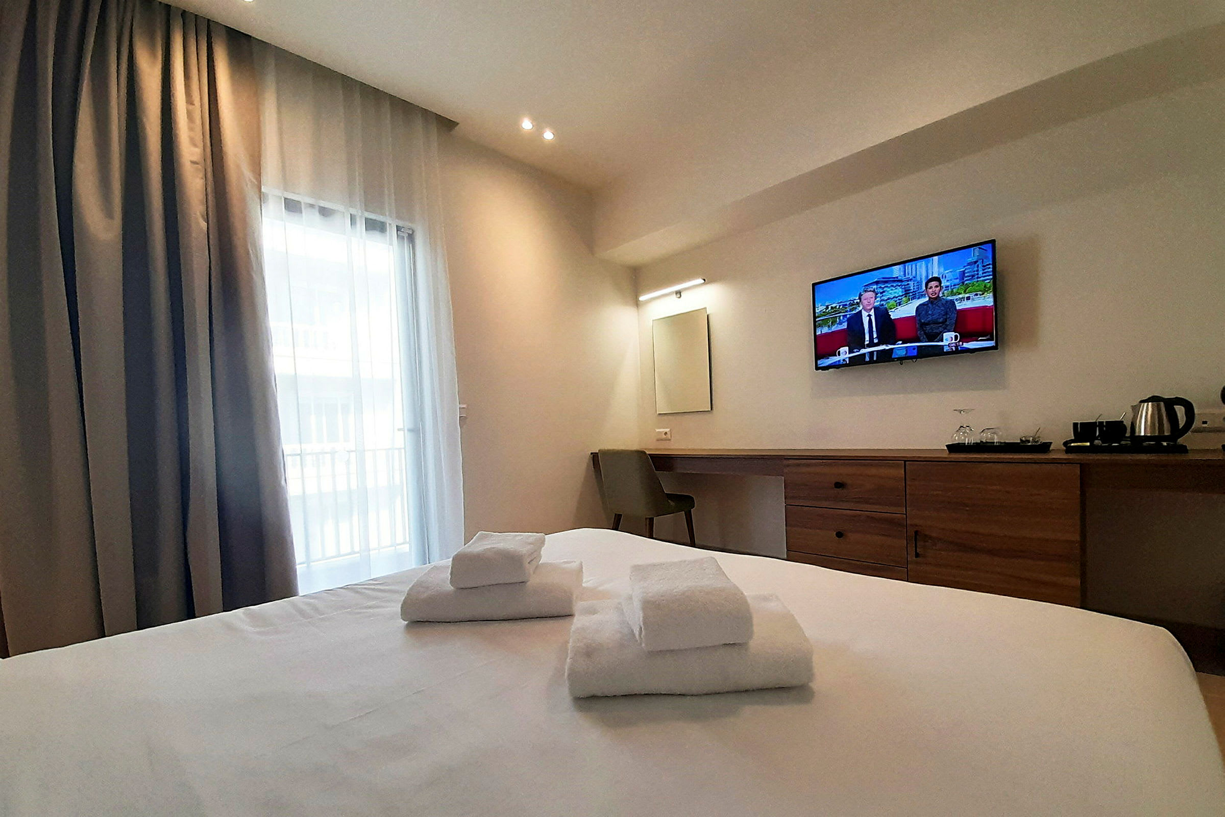 Et hotelværelse med en dobbeltseng, hvide håndklæder, et vægmonteret TV og et skrivebord med stol. Et vindue med gardiner ses. (AI-genereret tekst)