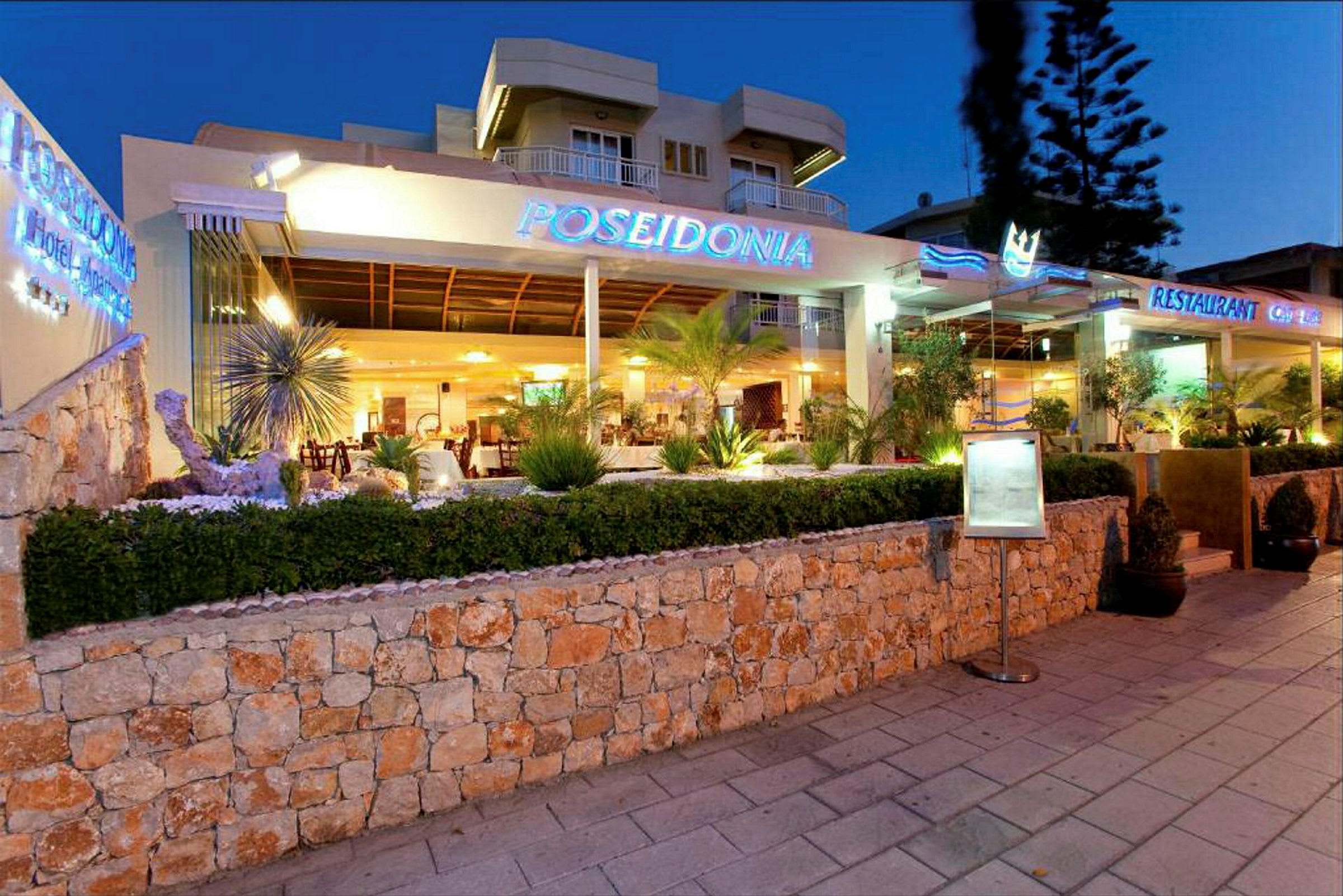 Hotel Poseidonia med aftenbelysning, omgivet af planter og et stenhegn. (AI-genereret tekst)