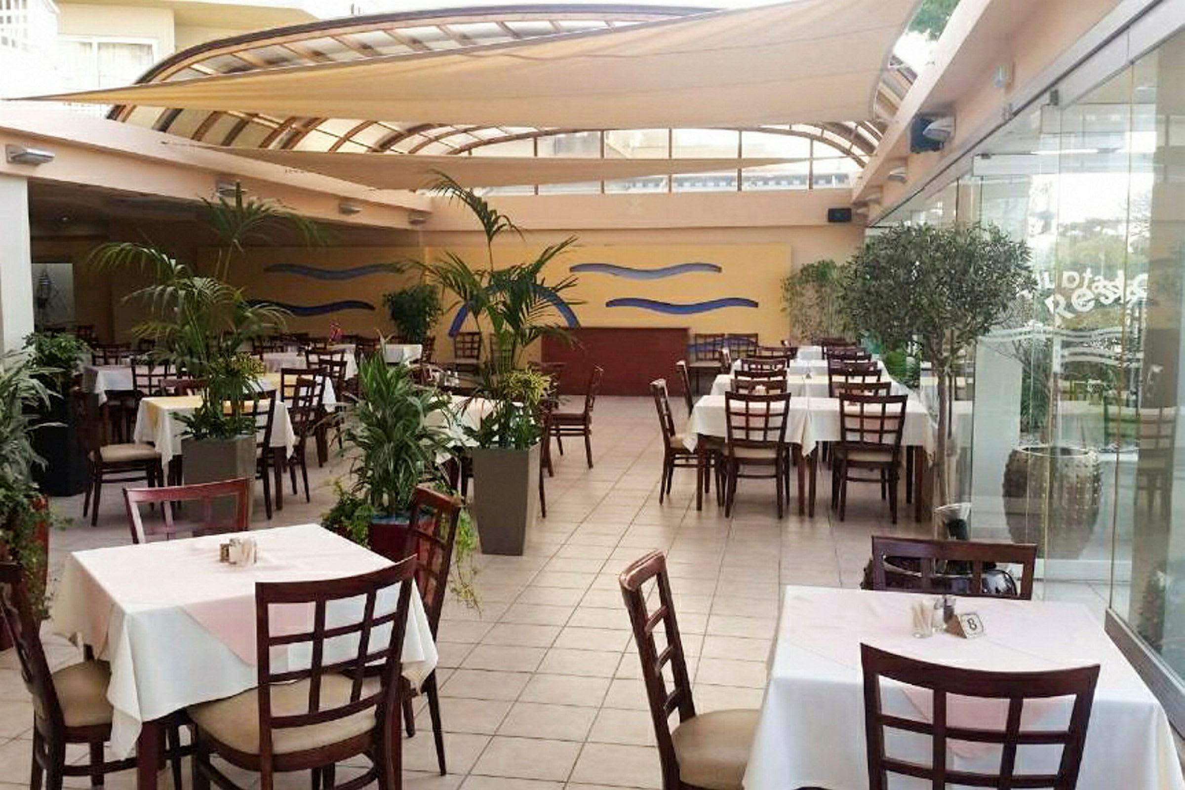 Billede af en restaurant med borde og stole omgivet af planter. (AI-genereret tekst)
