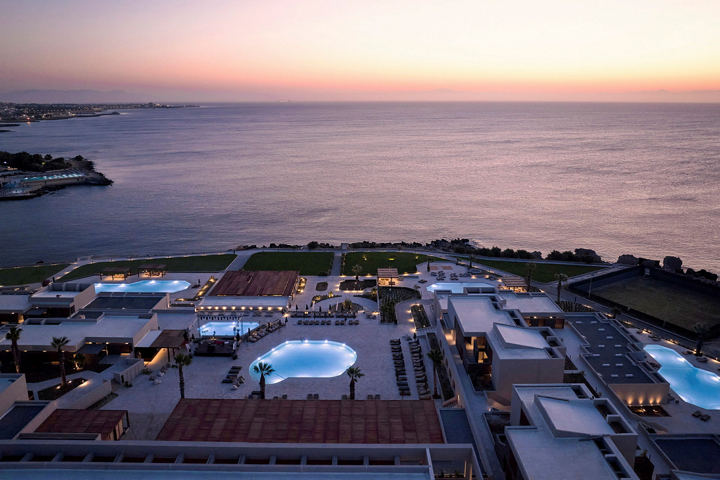 Luftfoto af hotelområde med pools ved havet i solnedgangen. (AI-genereret tekst)