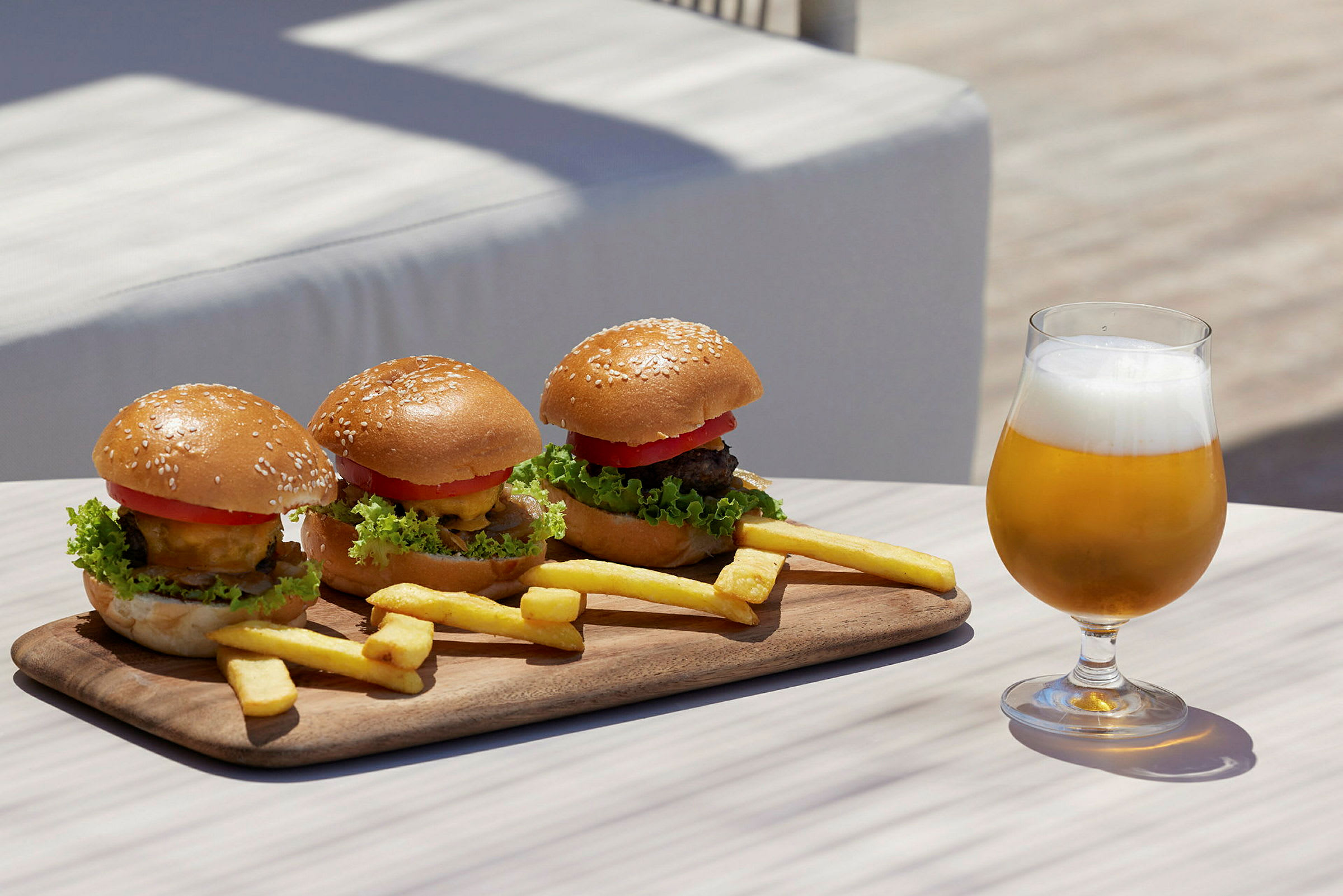 En træbakke med tre hamburgere og pommes frites samt et glas øl på et bord. (AI-genereret tekst)