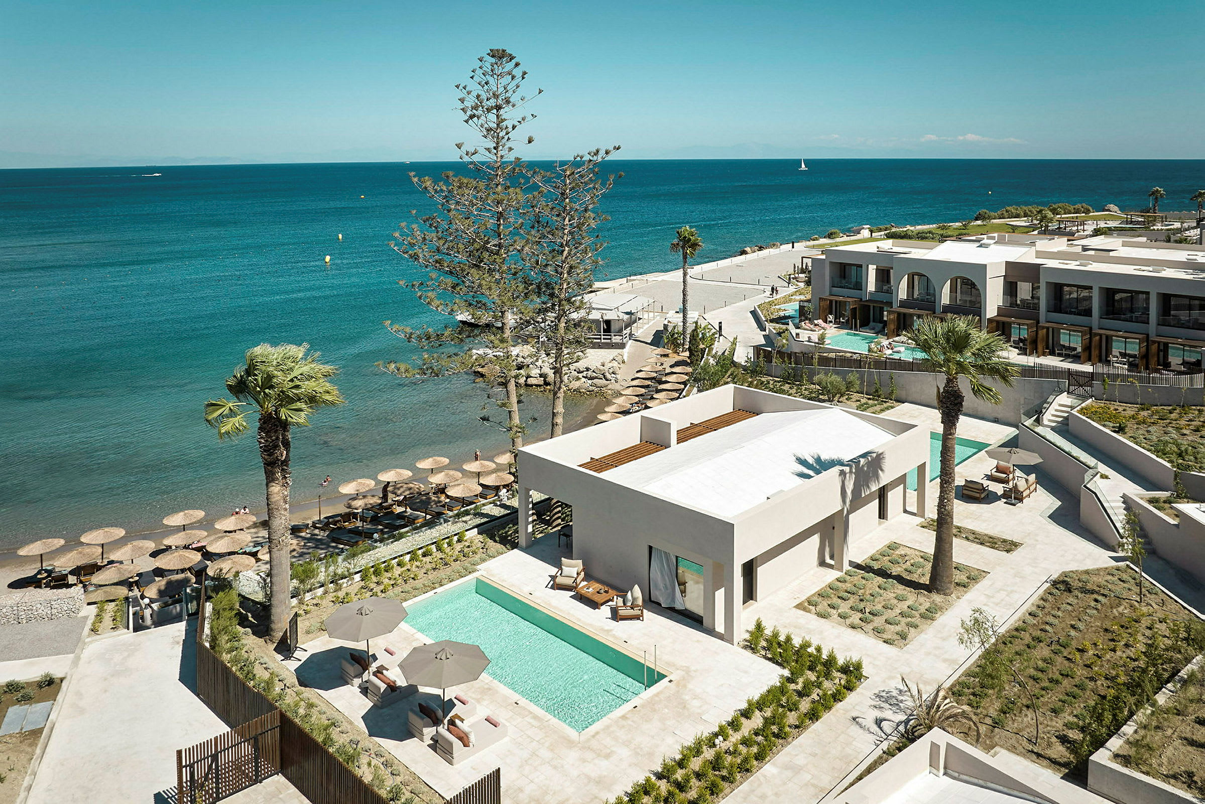 Udsigt over en strand med parasoller, et blåt hav og moderne bygninger med private swimmingpools. (AI-genereret tekst)