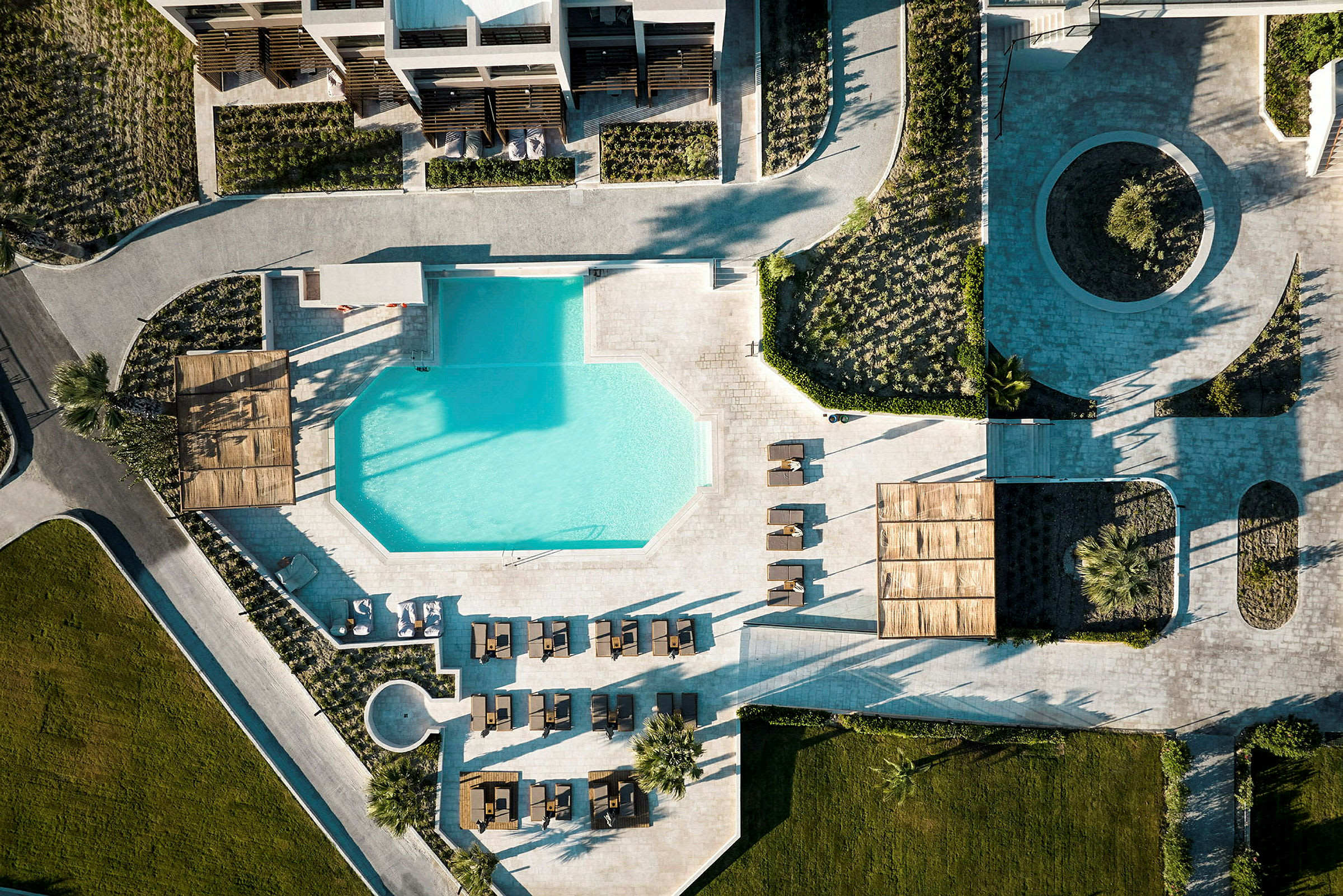 Luftfoto af en resort med en geometrisk formet pool omgivet af liggestole og grønne områder. (AI-genereret tekst)