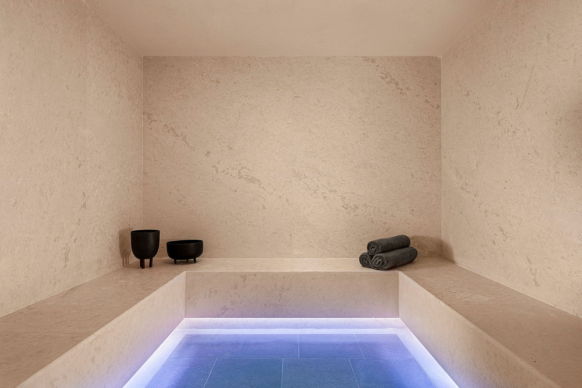 Et moderne spa-rum med minimalistisk indretning, bænksæde, grå oprullede håndklæder og to mørke dekorative skåle. (AI-genereret tekst)