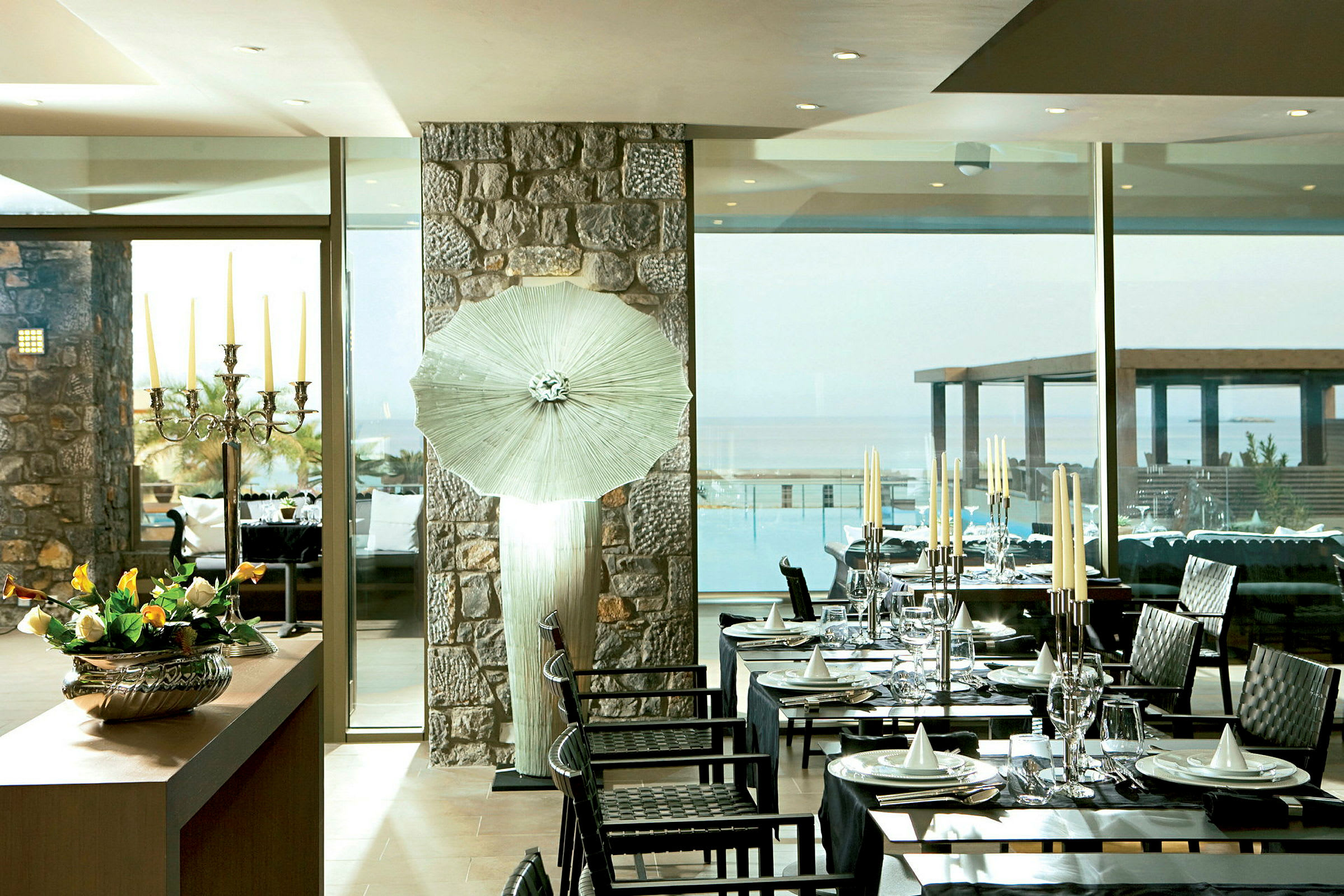En indendørsrestaurant med dækkede borde, en stor vinduesvæg med udsigt over en pool og havet samt en dekoreret lysestage og blomster på et bord. (AI-genereret tekst)