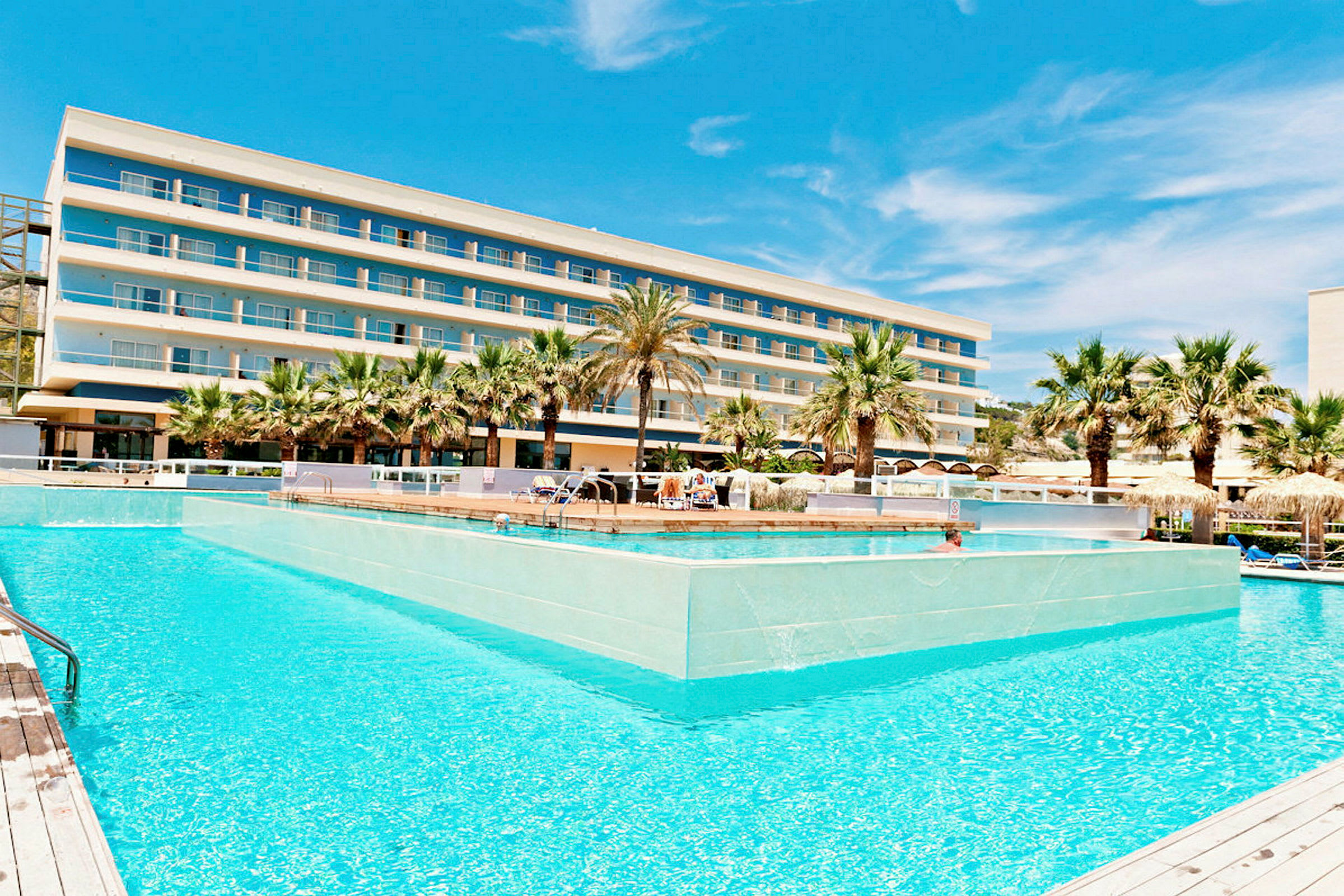 Et hotel med flere etager bag en stor pool omgivet af palmer. (AI-genereret tekst)