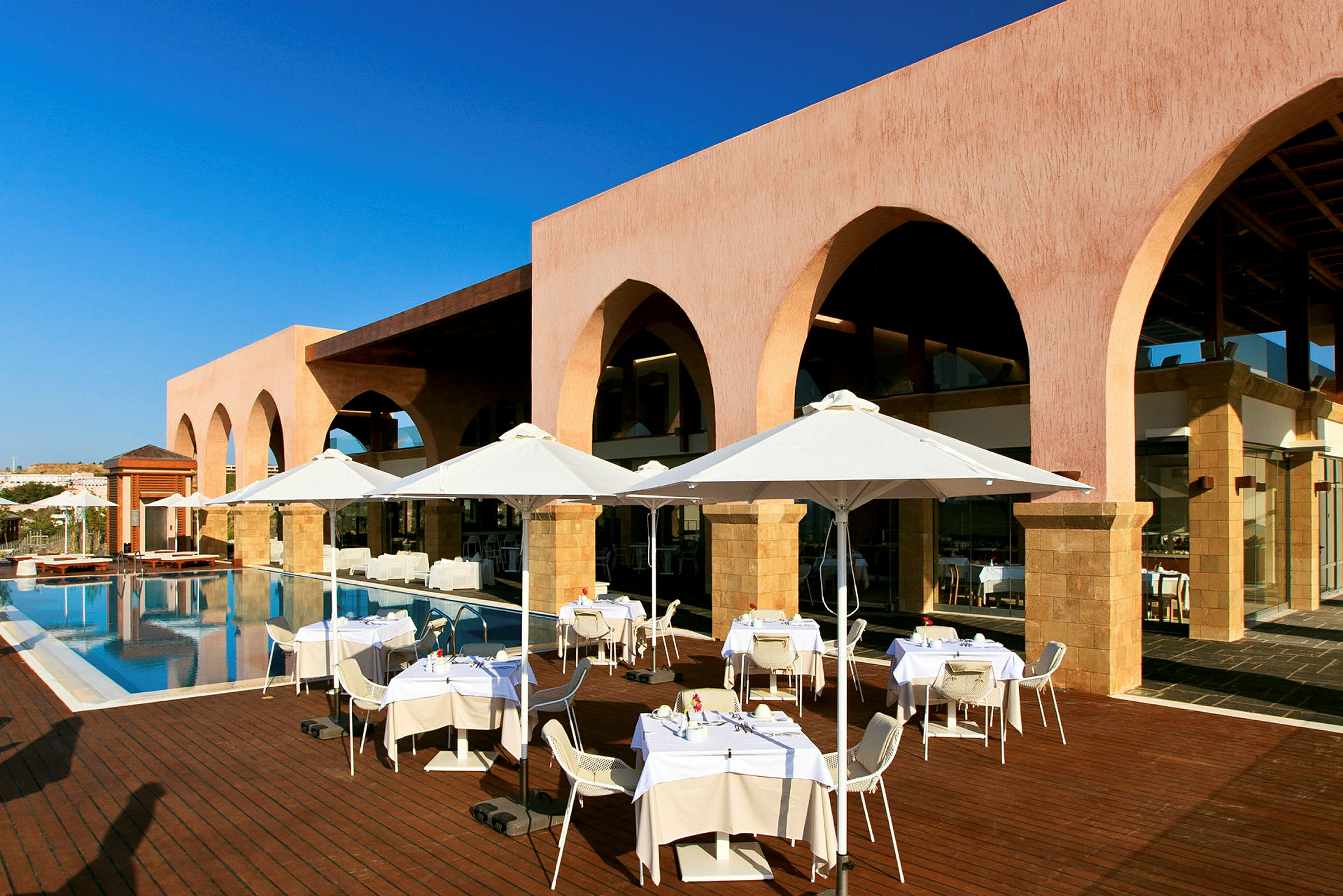 En udendørsrestaurant med hvide parasoller ved en pool foran en bygning med buer. (AI-genereret tekst)