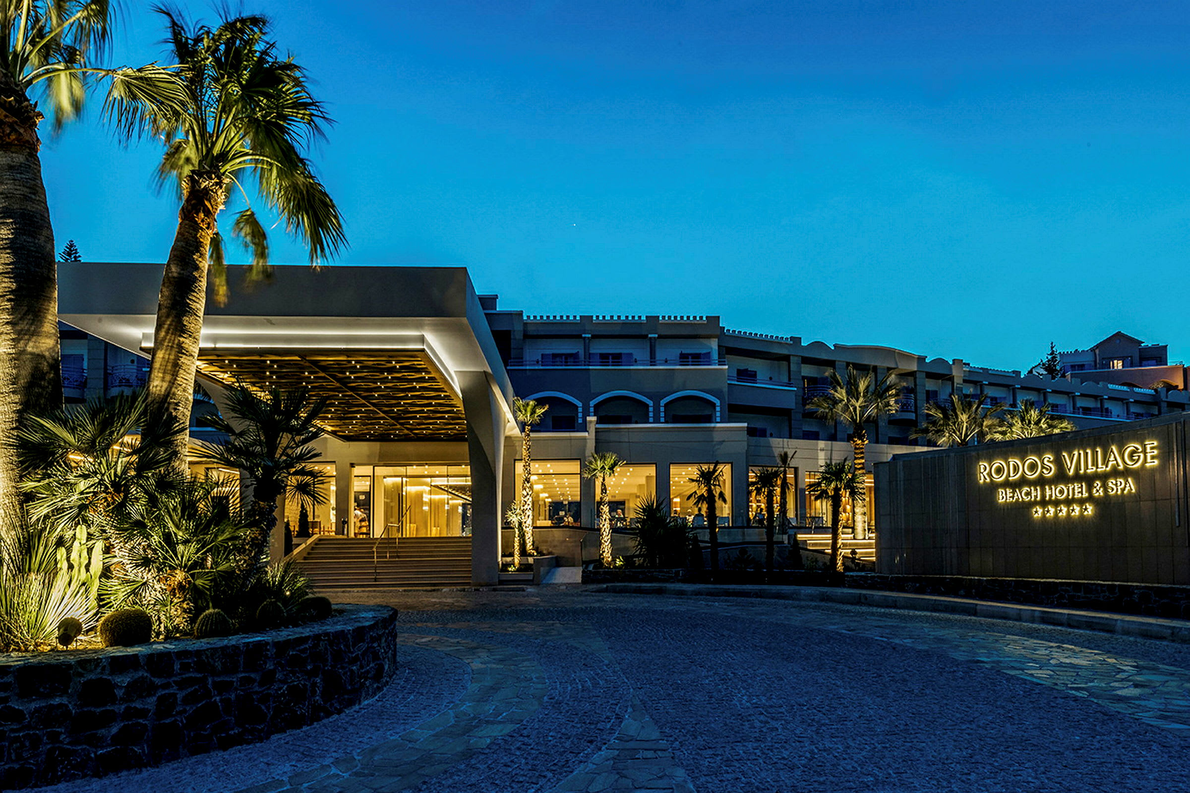 Et hotel om aftenen med oplyst indgang og skilt med teksten 'Rodos Village Beach Hotel & Spa'. (AI-genereret tekst)