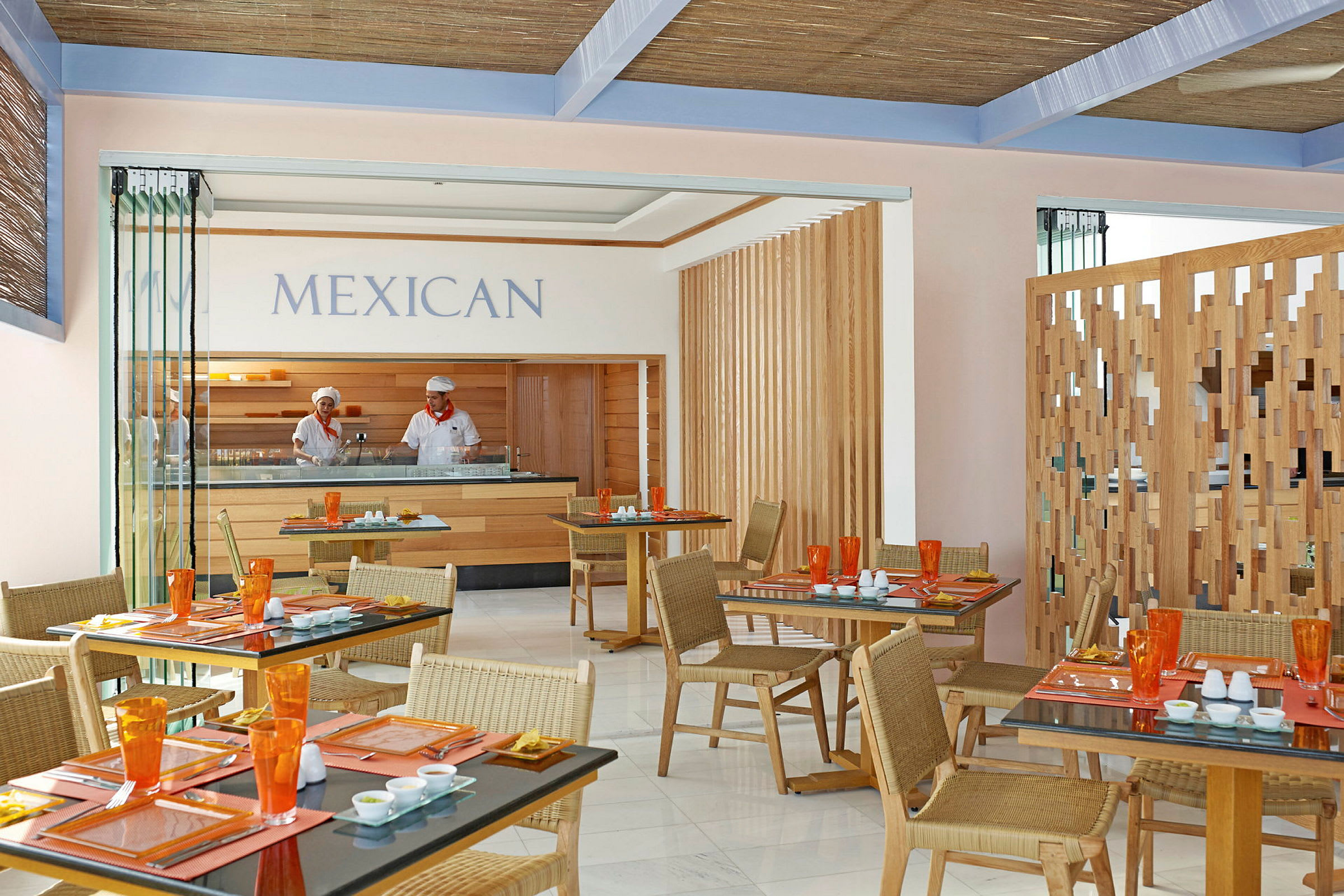 En hyggelig restaurant med mexicansk indretning viser borde dækket med farverige glas og tallerkener, mens en serveringsdisk og del af køkkenet er synlig i baggrunden. (AI-genereret tekst)