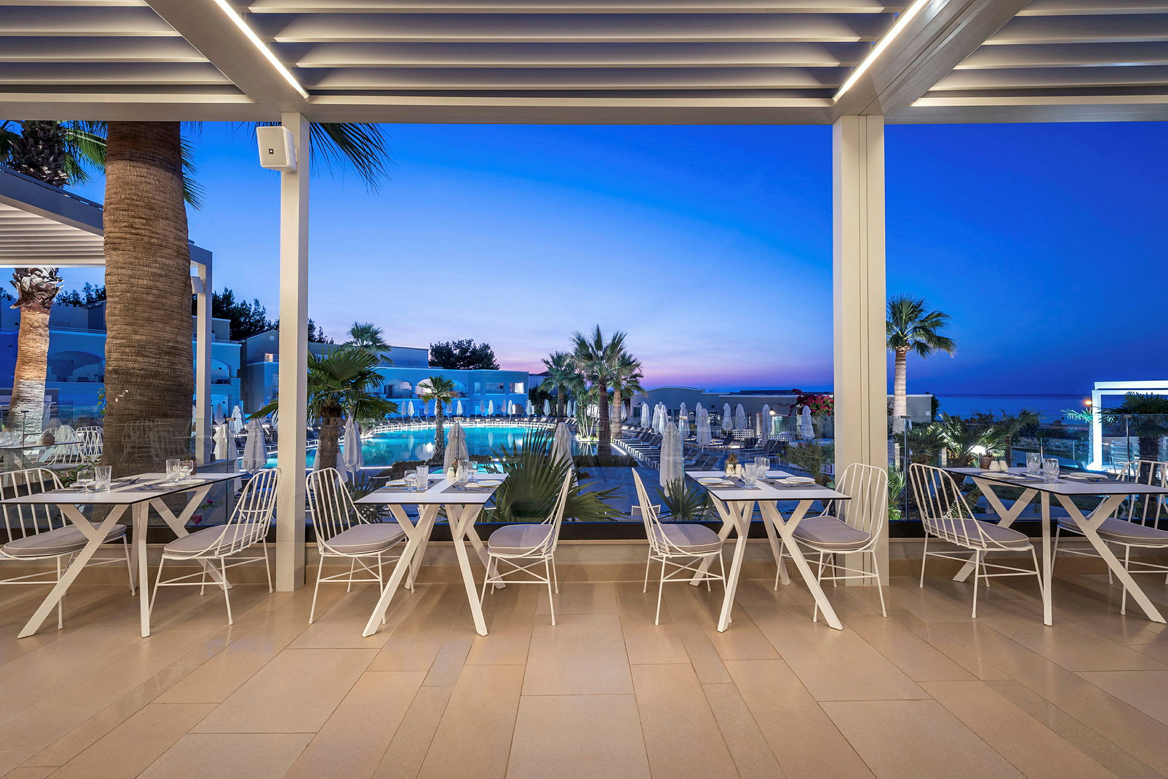 En udendørsrestaurant med hvidt interiør og udsigt over en pool omgivet af palmer under en klar tusmørkehimmel. (AI-genereret tekst)
