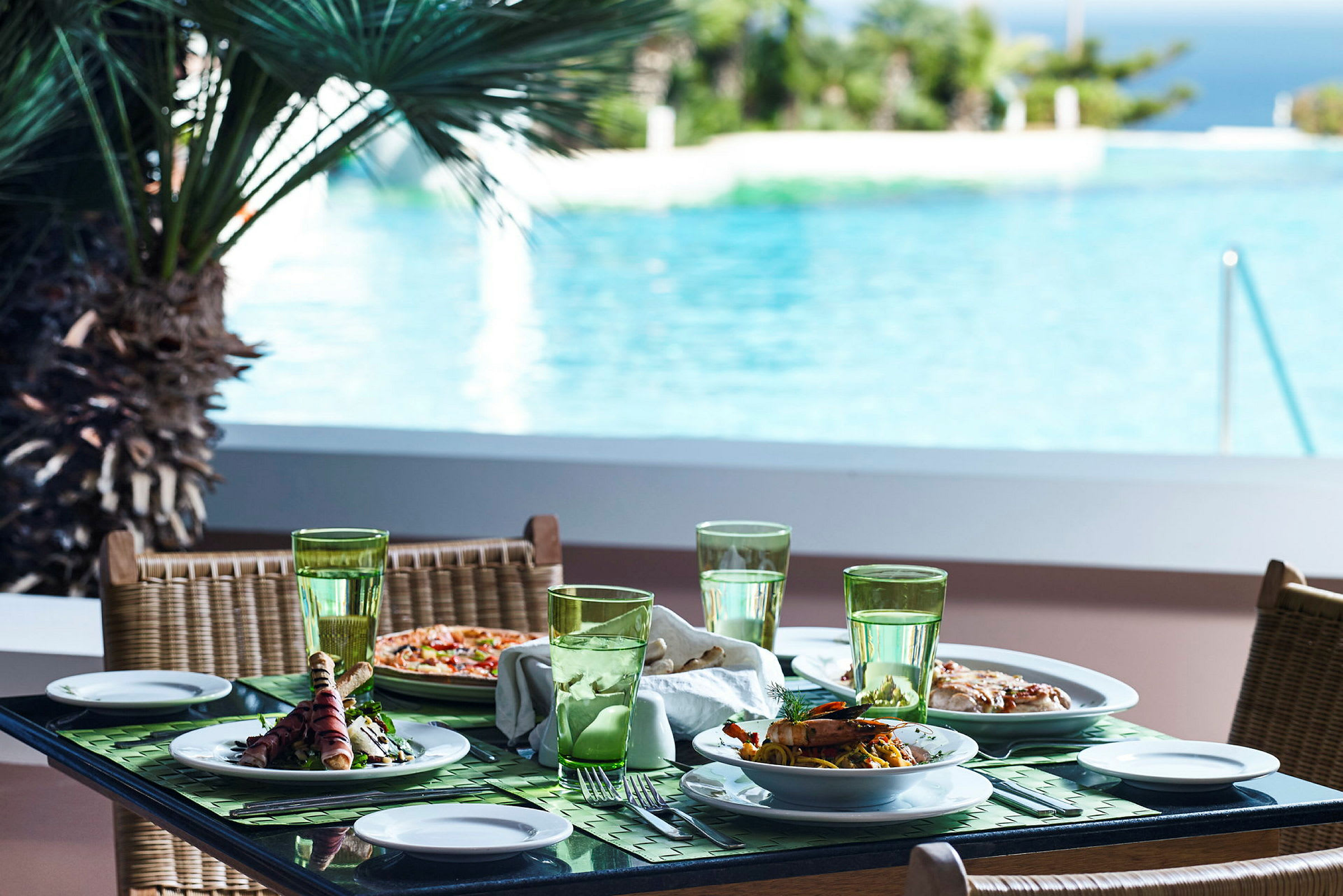 Et dækket bord med retter og glas med udsigt over en swimmingpool og omgivende grønne områder. (AI-genereret tekst)
