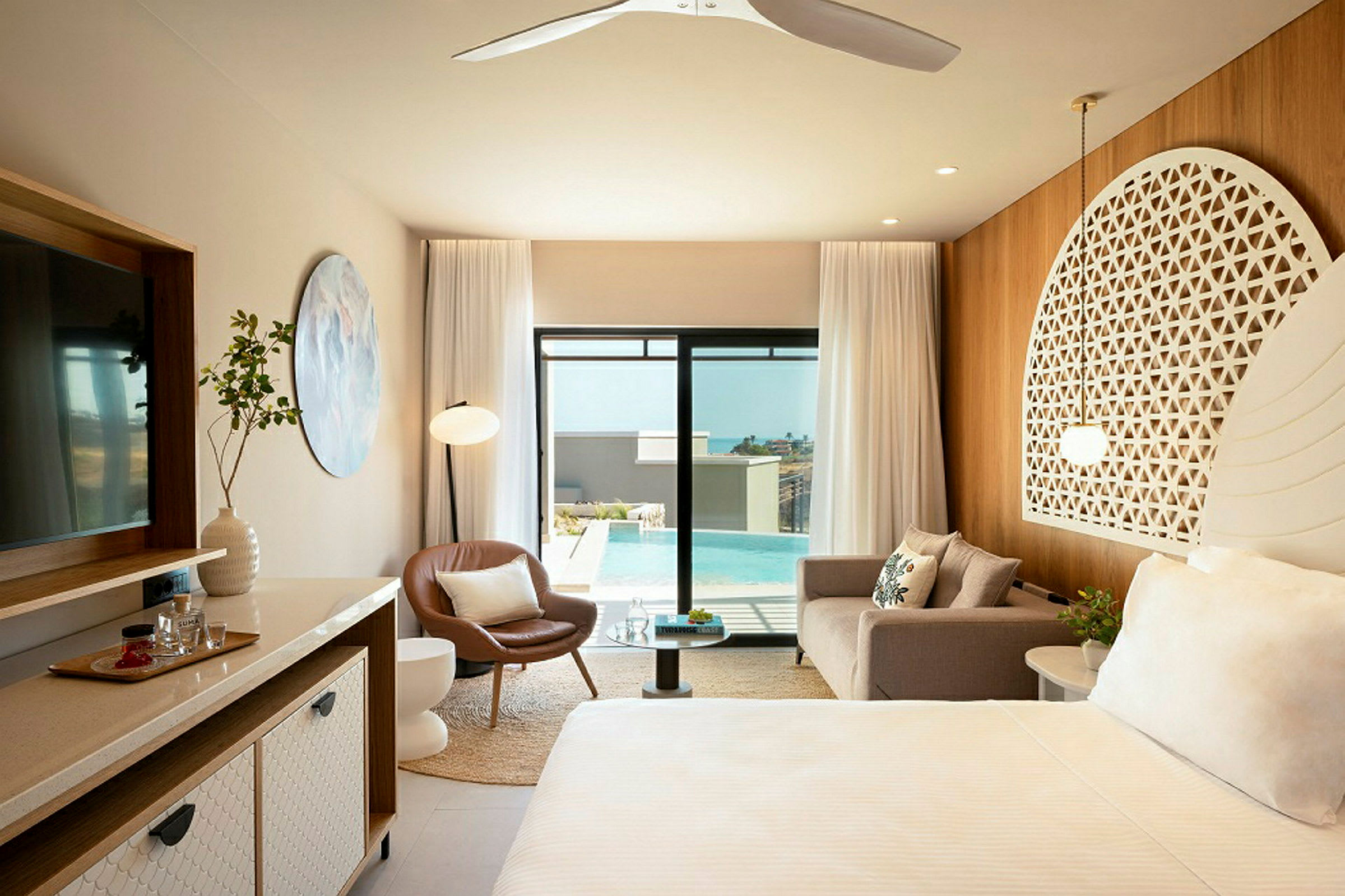 Et elegant hotelværelse med moderne møbler, en sofa, en lænestol, et sengeområde med dekorativ hovedgærde, et stort spejl og glasdøre, der åbner ud til en udendørs swimmingpool. (AI-genereret tekst)