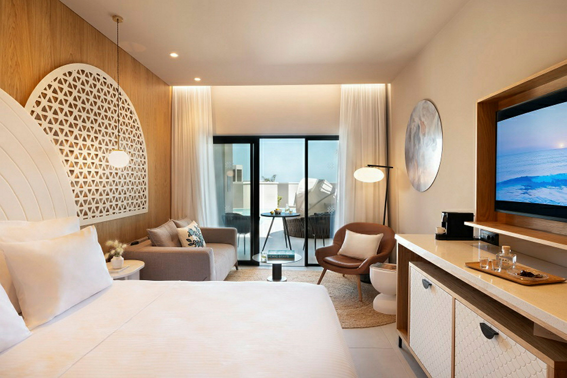 Et moderne hotelværelse med lyse møbler og dekorationer, herunder en seng, sofa, stol og TV. Værelset har store glasdøre, der fører ud til en balkon. (AI-genereret tekst)