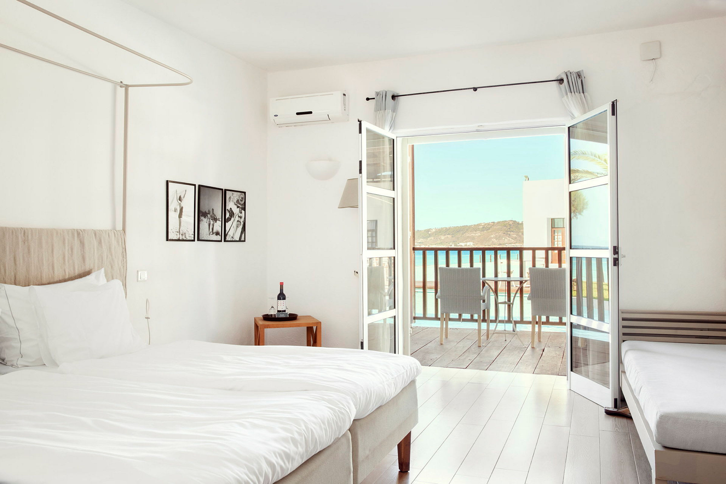 Et lyst hotelværelse med en seng, små møbler og en balkon med udsigt over havet. (AI-genereret tekst)