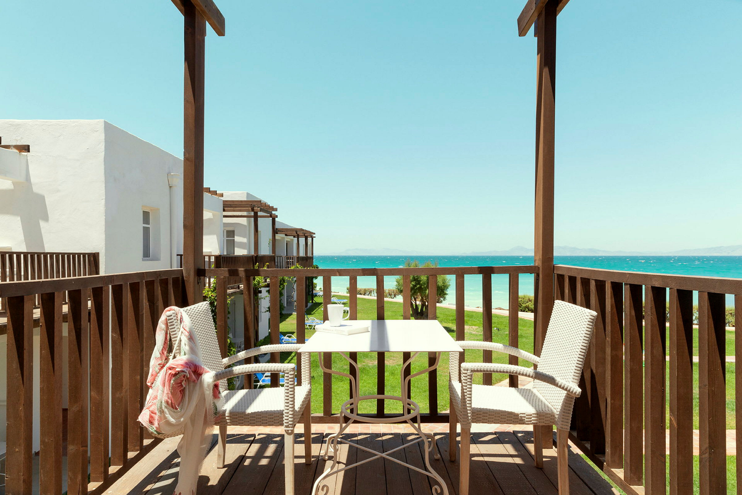Udsigt fra en balkon med bord og stole mod havet og bygninger. (AI-genereret tekst)