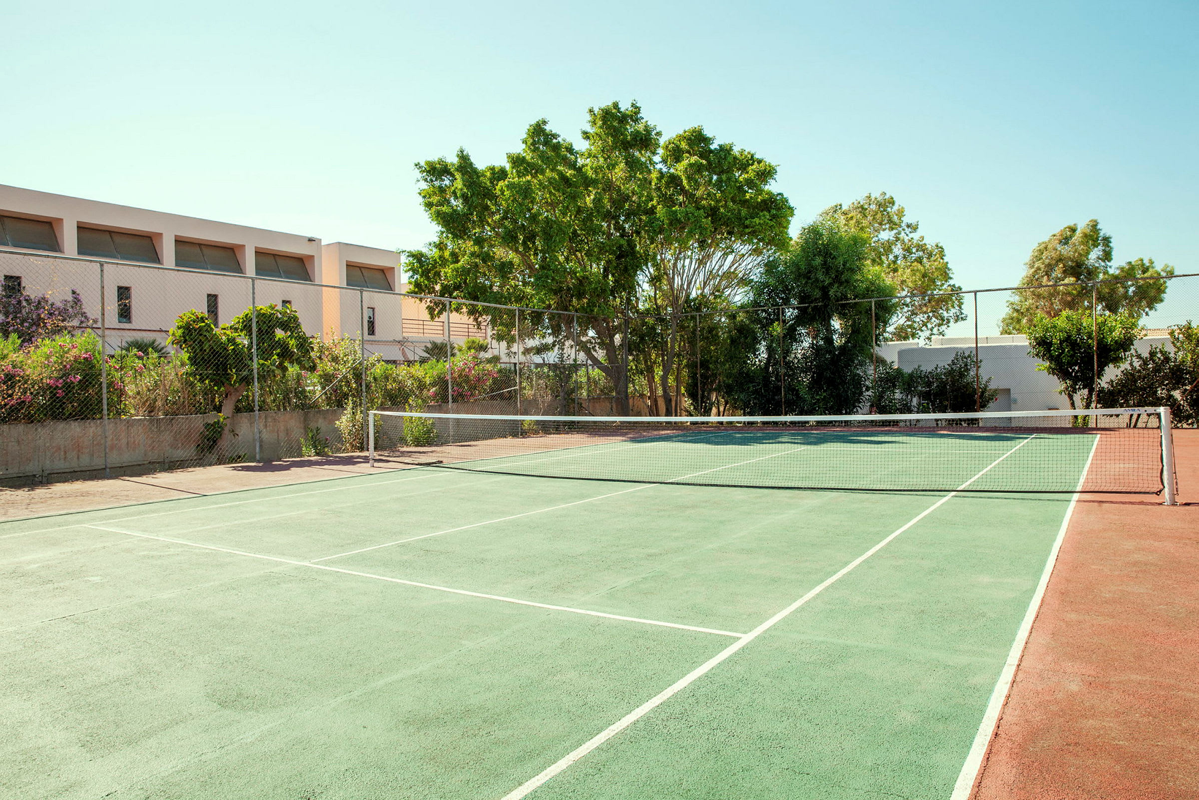 En tennisbane med grøn overflade og net, omgivet af træer og bygninger. (AI-genereret tekst)