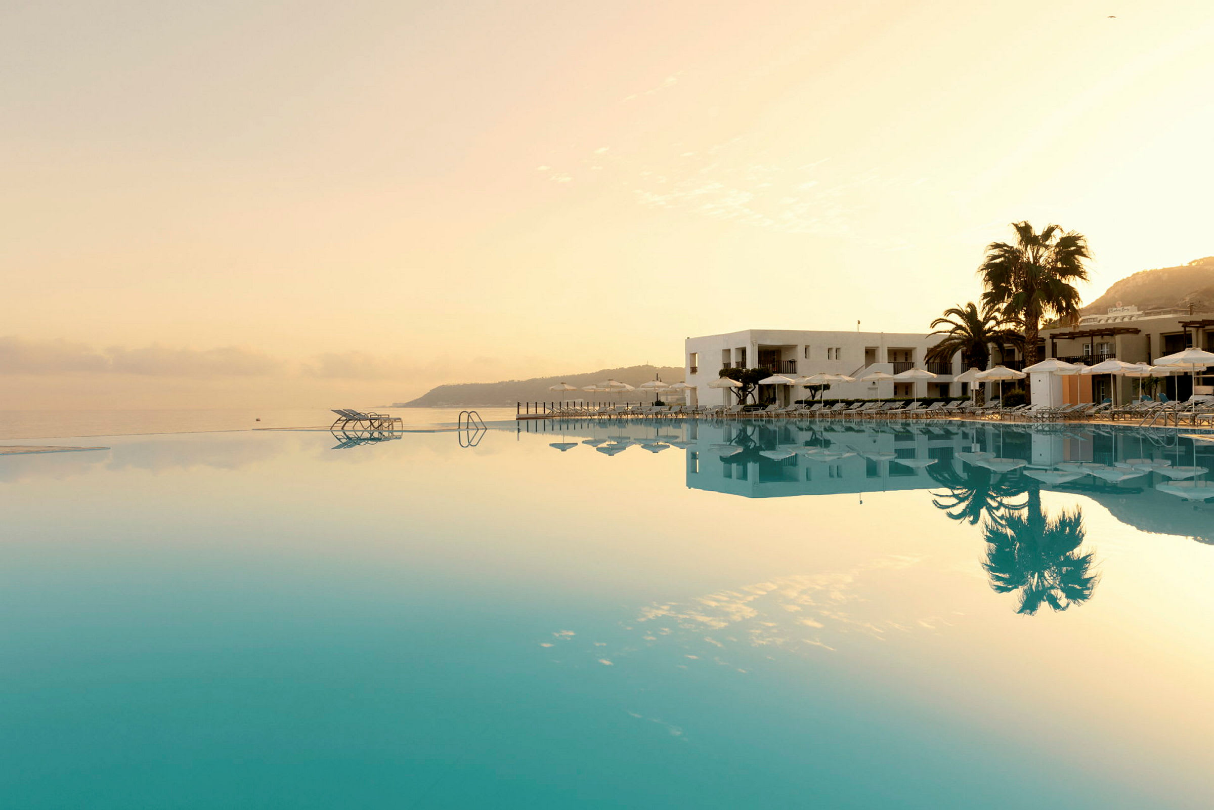 Billedet viser en infinitypool ved siden af et hotel med liggestole og parasoller, omgivet af palmer og morgenlys. (AI-genereret tekst)