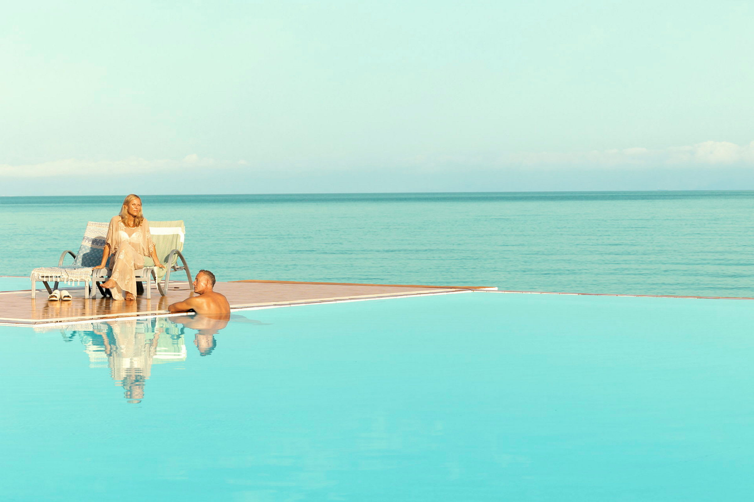 En infinitypool med udsigt over havet, med en person ved poolkanten og en anden på en solseng. (AI-genereret tekst)