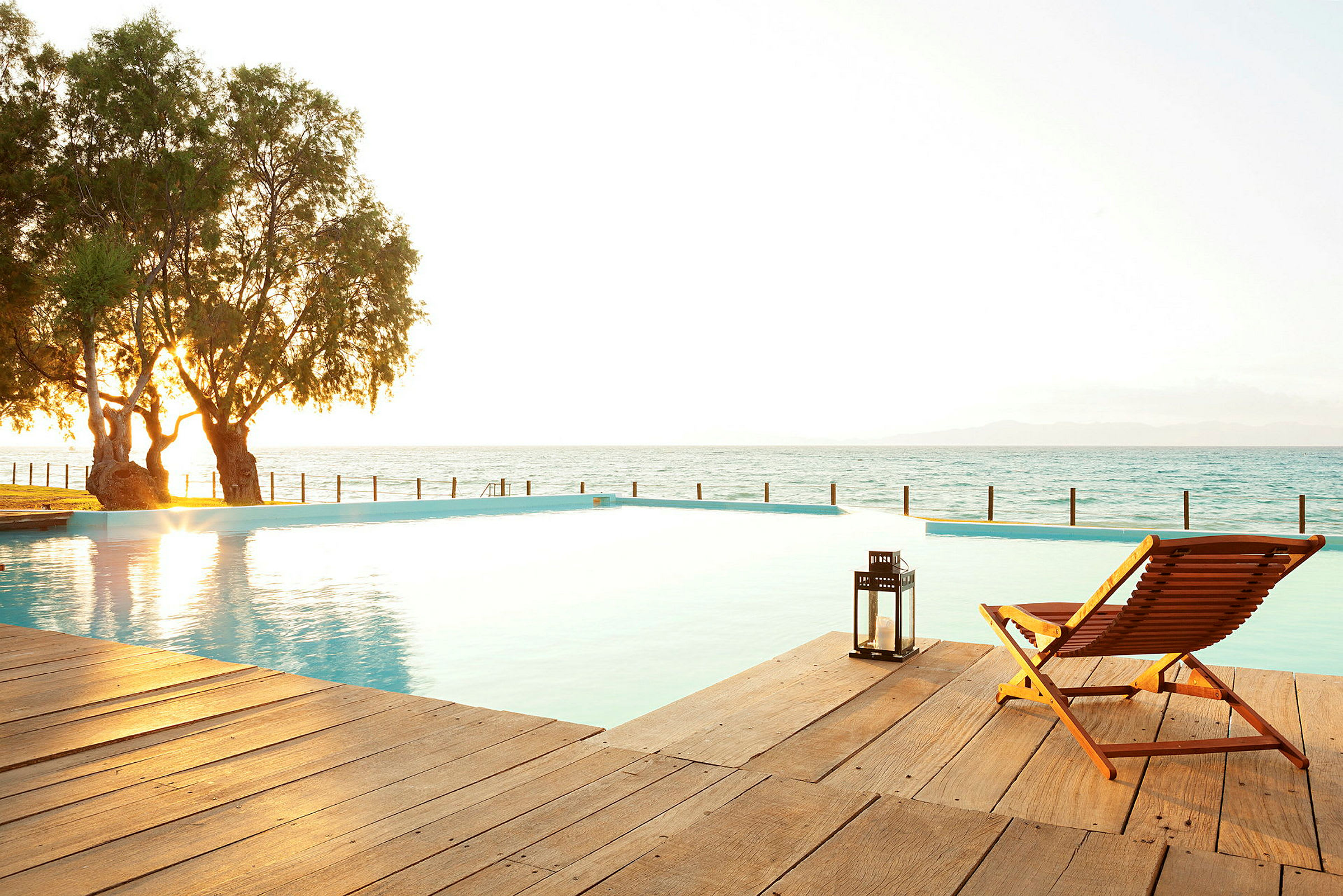 Fotografi af en infinity-pool ved havet i solnedgang, med en træstol og en lanterne på en træterrasse. (AI-genereret tekst)