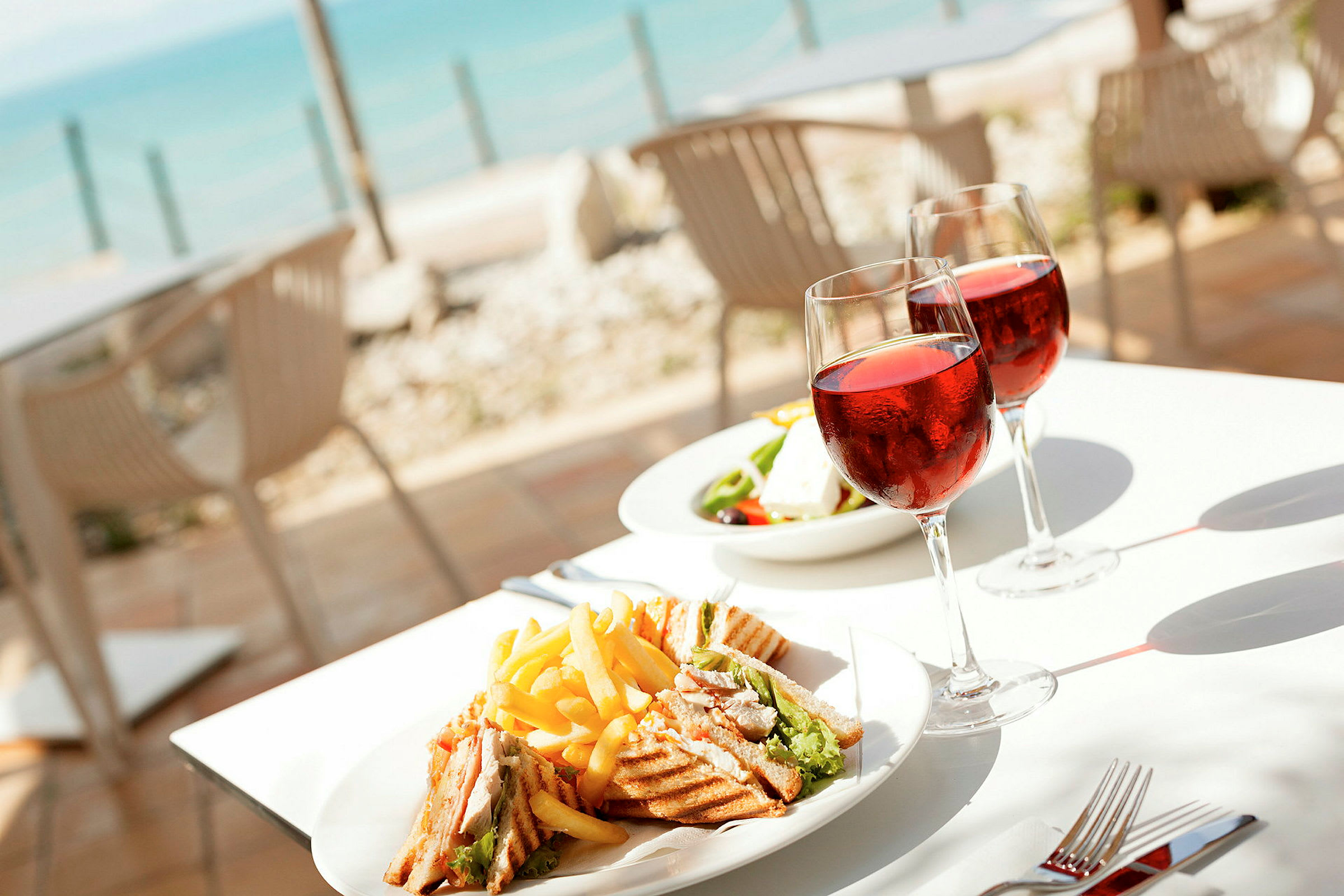 Tallerken med sandwich og pommes frites samt to glas vin ved et bord nær havet. (AI-genereret tekst)