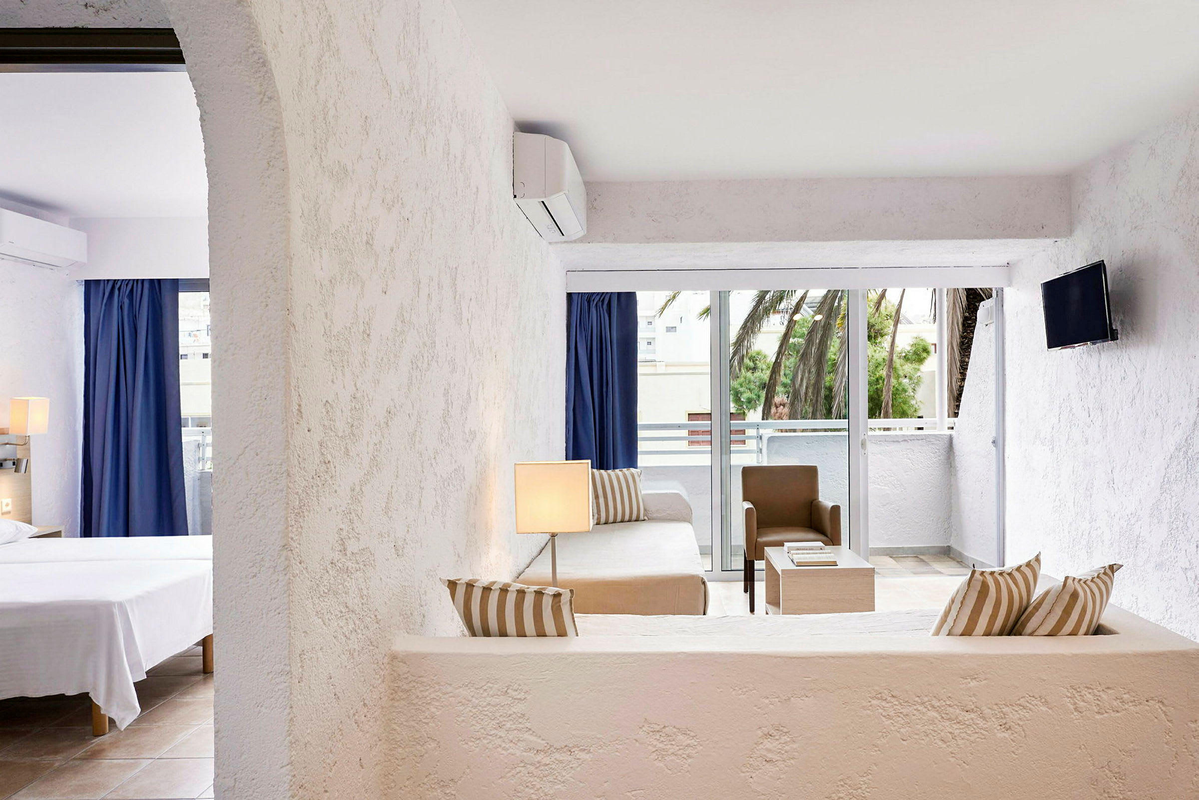 Et lyst hotelværelse med en stueafdeling, møbler som sofa, bord og stol i neutrale farver, og et åbent soveværelse. Der er en balkon med udsigt mod palmer. (AI-genereret tekst)