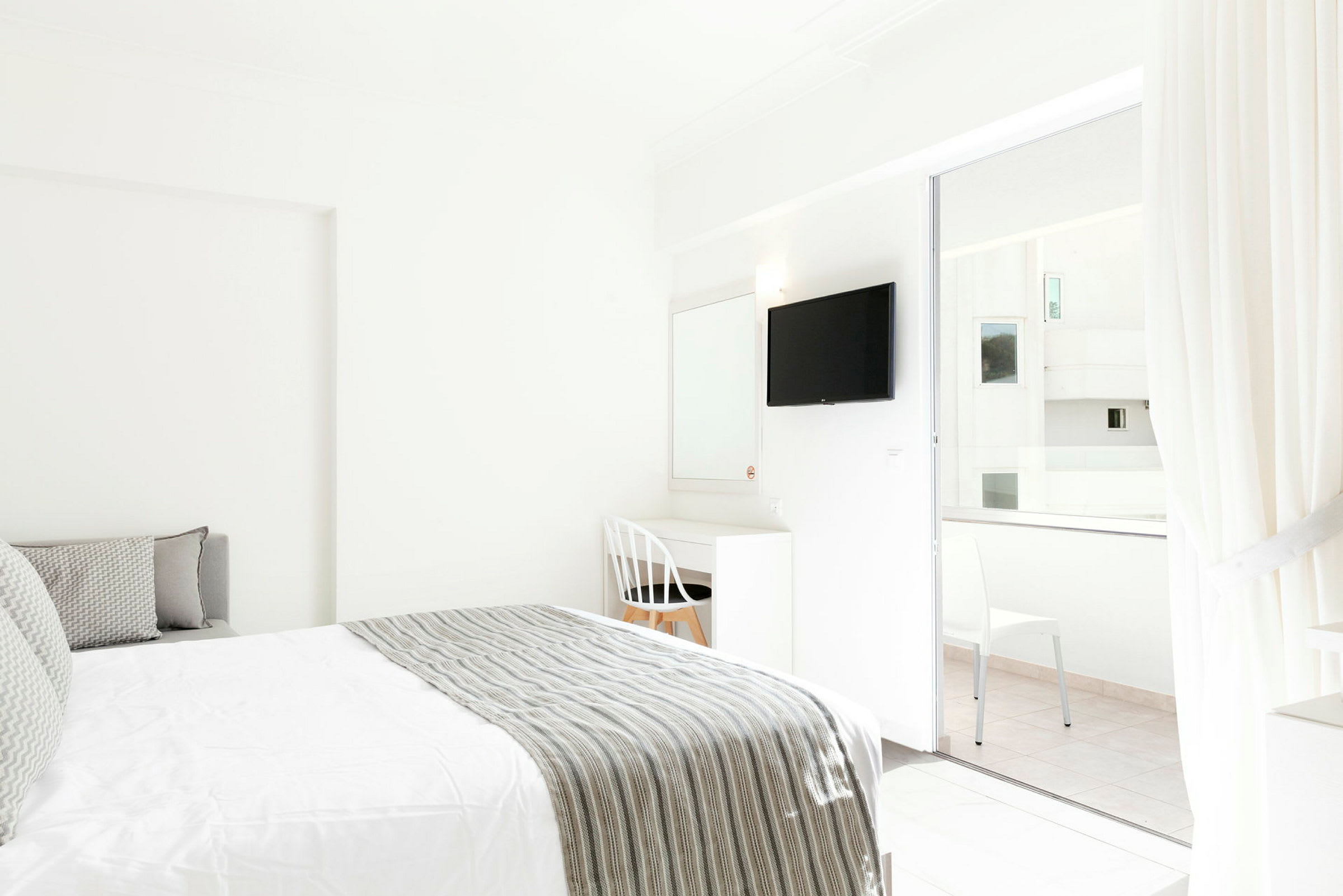 Interiøret i et lyst hotelværelse med en seng og skrivebord ved siden af en hvid stol. Et TV er monteret på væggen, og glasdøre fører til en balkon. (AI-genereret tekst)
