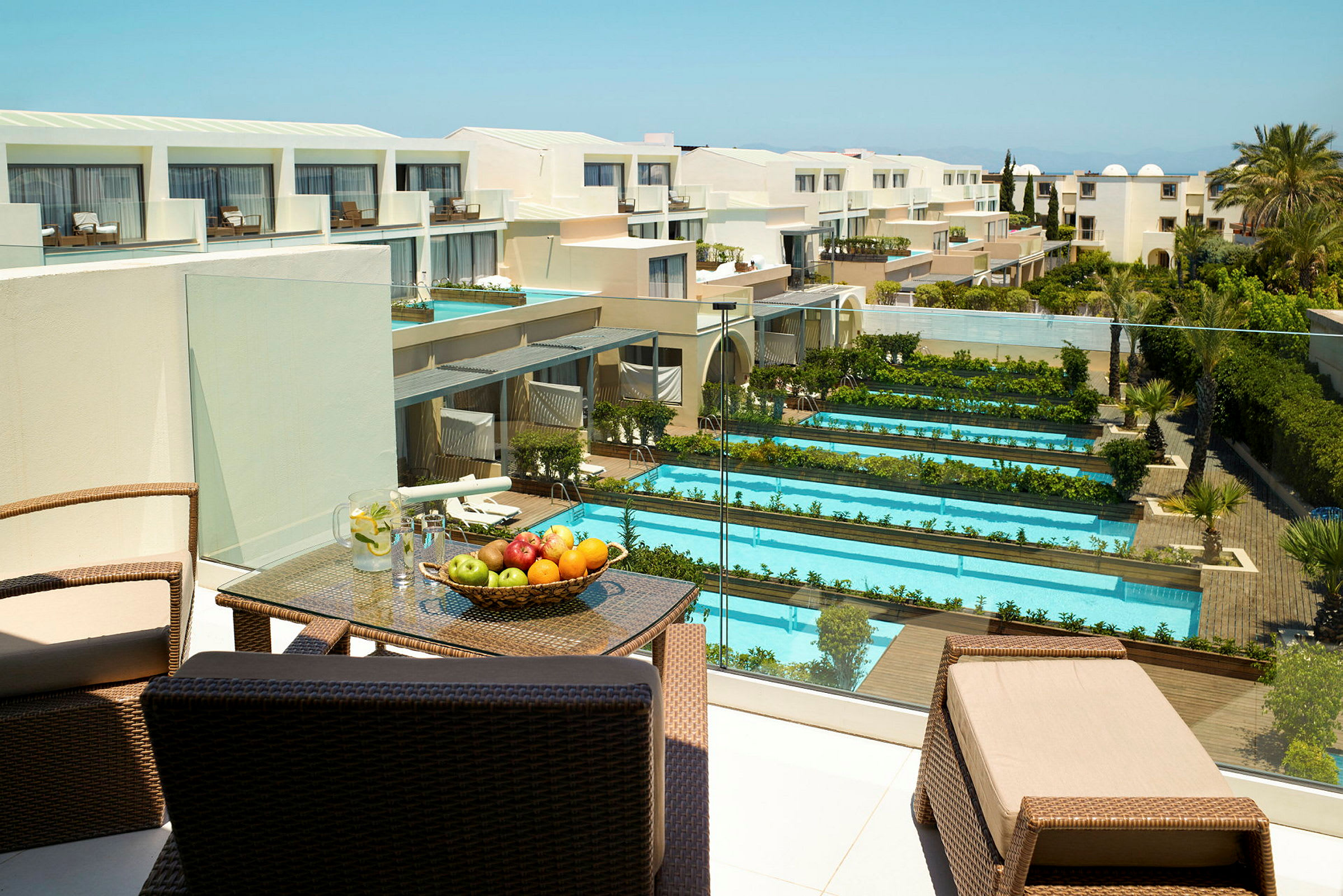 Hotelområde med flere pooler og grønne områder, samt en terrasse med møbler og frugt. Udsigt over en moderne bygning. (AI-genereret tekst)