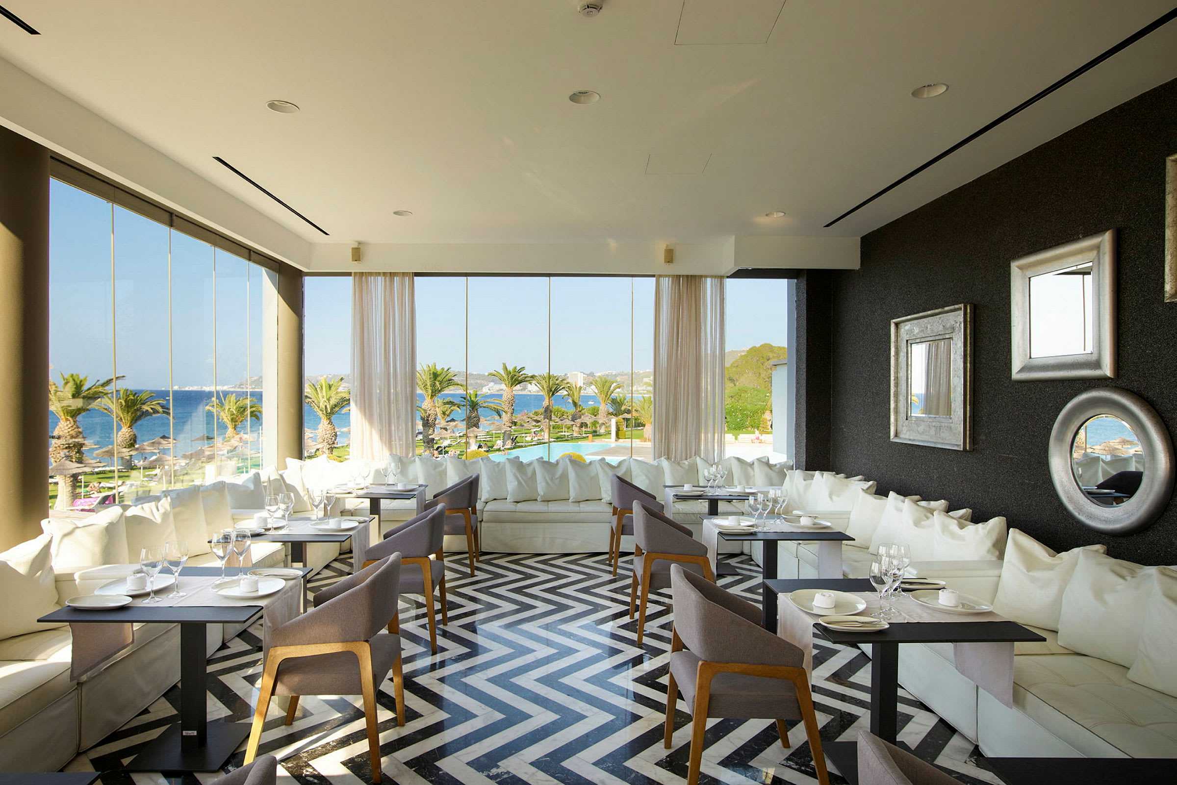 Interiør af en restaurant med store vinduer, moderne møbler og udsigt over palmer og havet. (AI-genereret tekst)